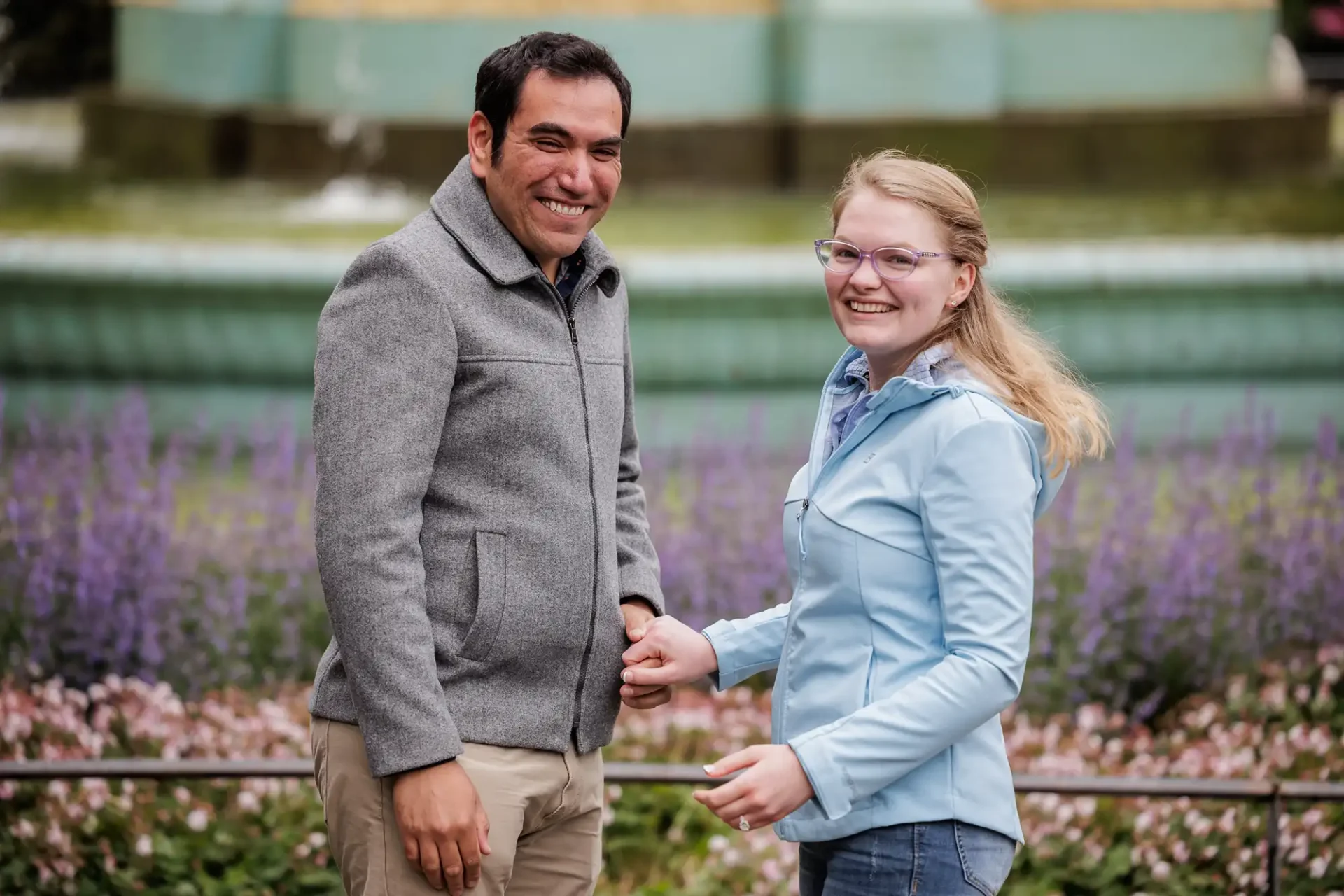 A man and a woman stand outdoors holding hands, smiling at the camera, with purple flowers and a fountain in the background.