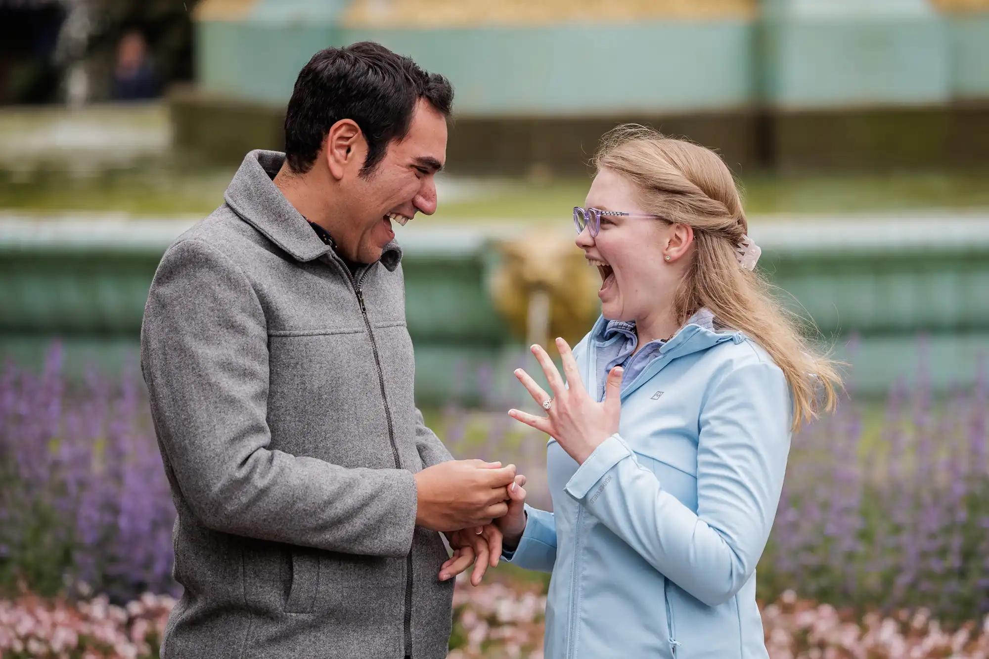 A man and woman stand outdoors; the woman looks surprised and shows off a ring on her hand, while the man smiles and holds her other hand.