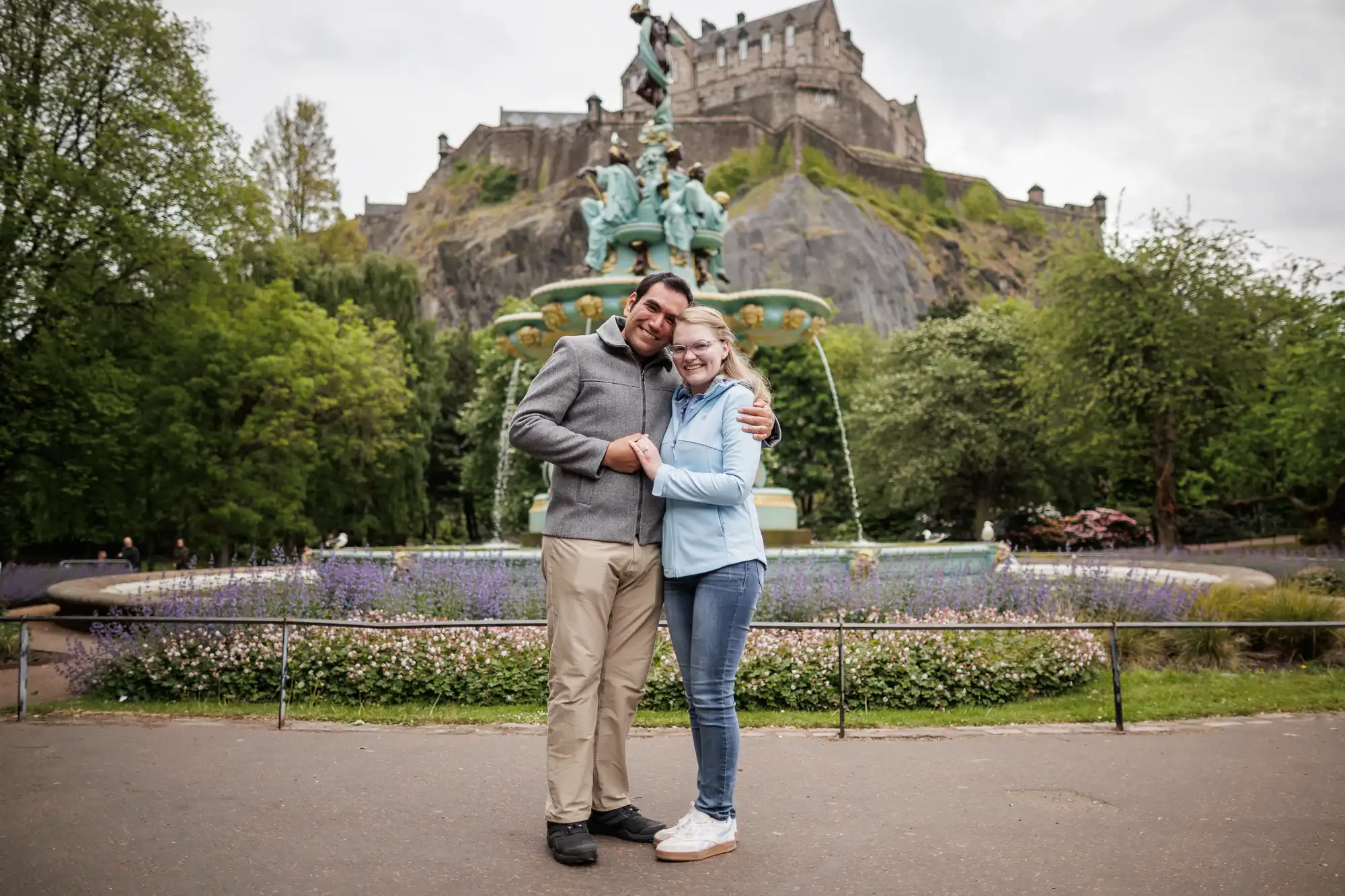 Two people stand smiling and embracing in front of a decorative fountain, with a historic castle on a hill in the background.