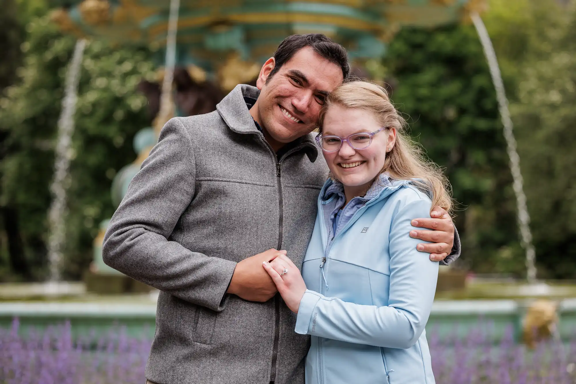 A man and woman stand together outdoors, smiling and embracing in front of a fountain and greenery. The woman displays a ring on her finger.