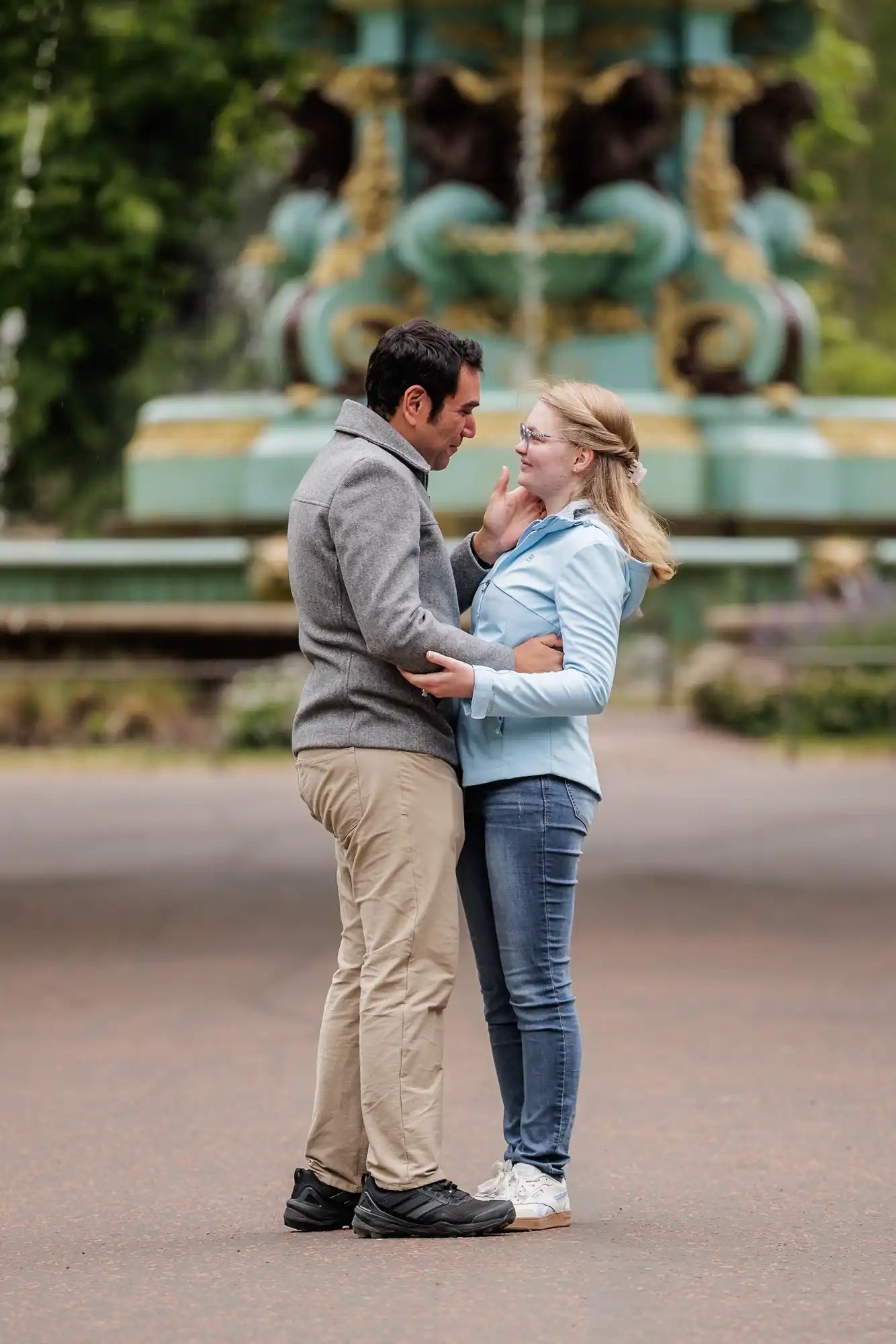 A man and woman stand facing each other outdoors, smiling and embracing, with a decorative fountain in the background.