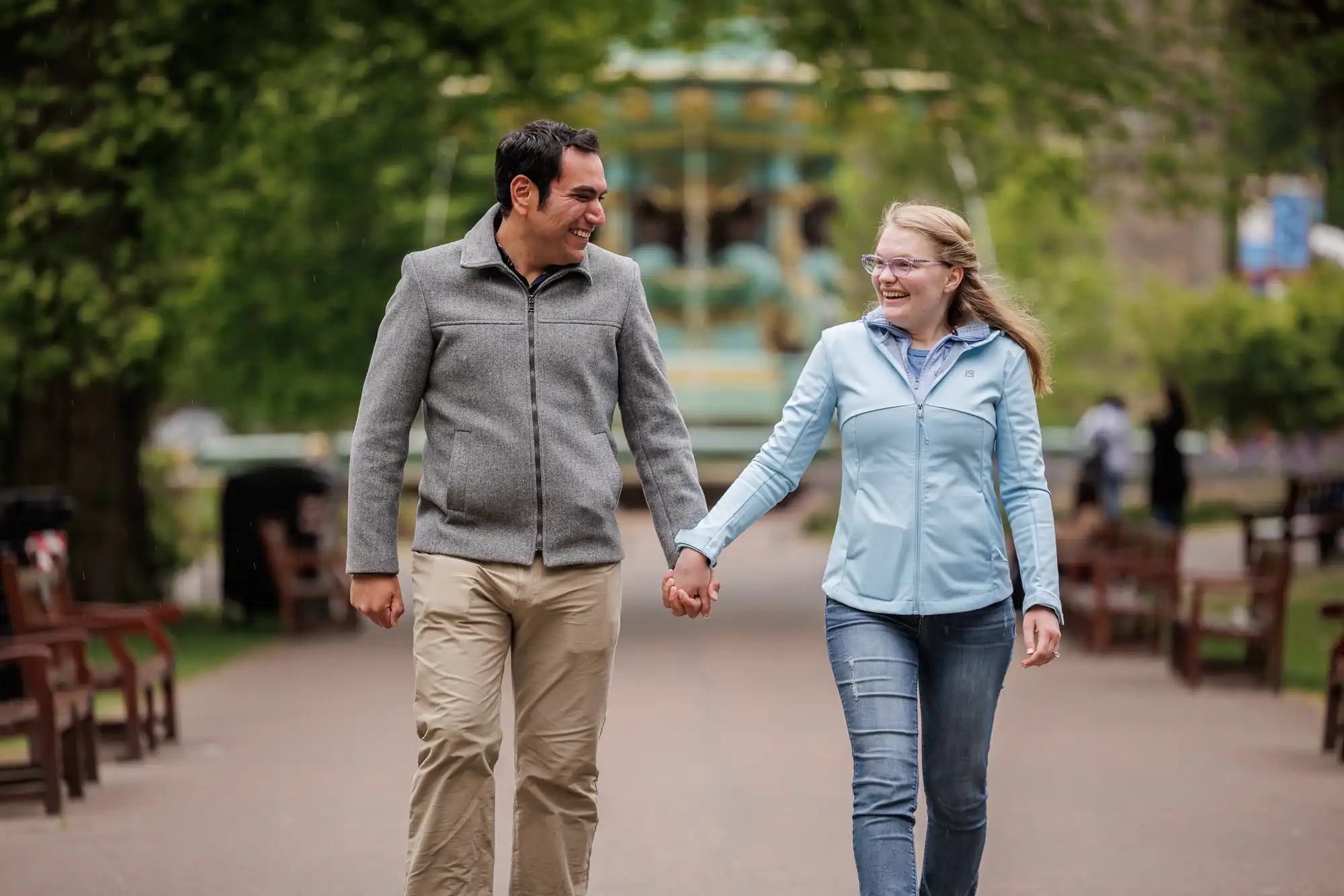 A man and a woman holding hands and smiling while walking down a tree-lined park path.