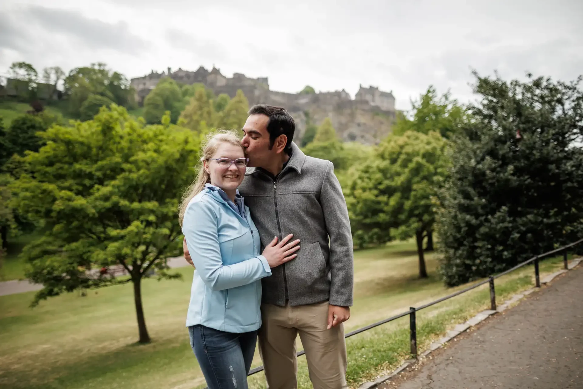 A man kisses a woman on the temple as they pose together in a park with trees and a distant castle in the background.