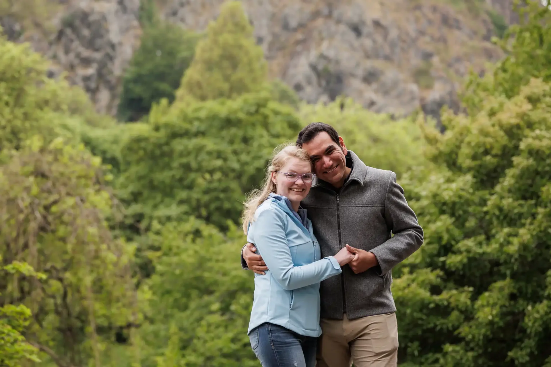 Two people stand close together, smiling and holding hands, with a backdrop of green trees and rocky hills.
