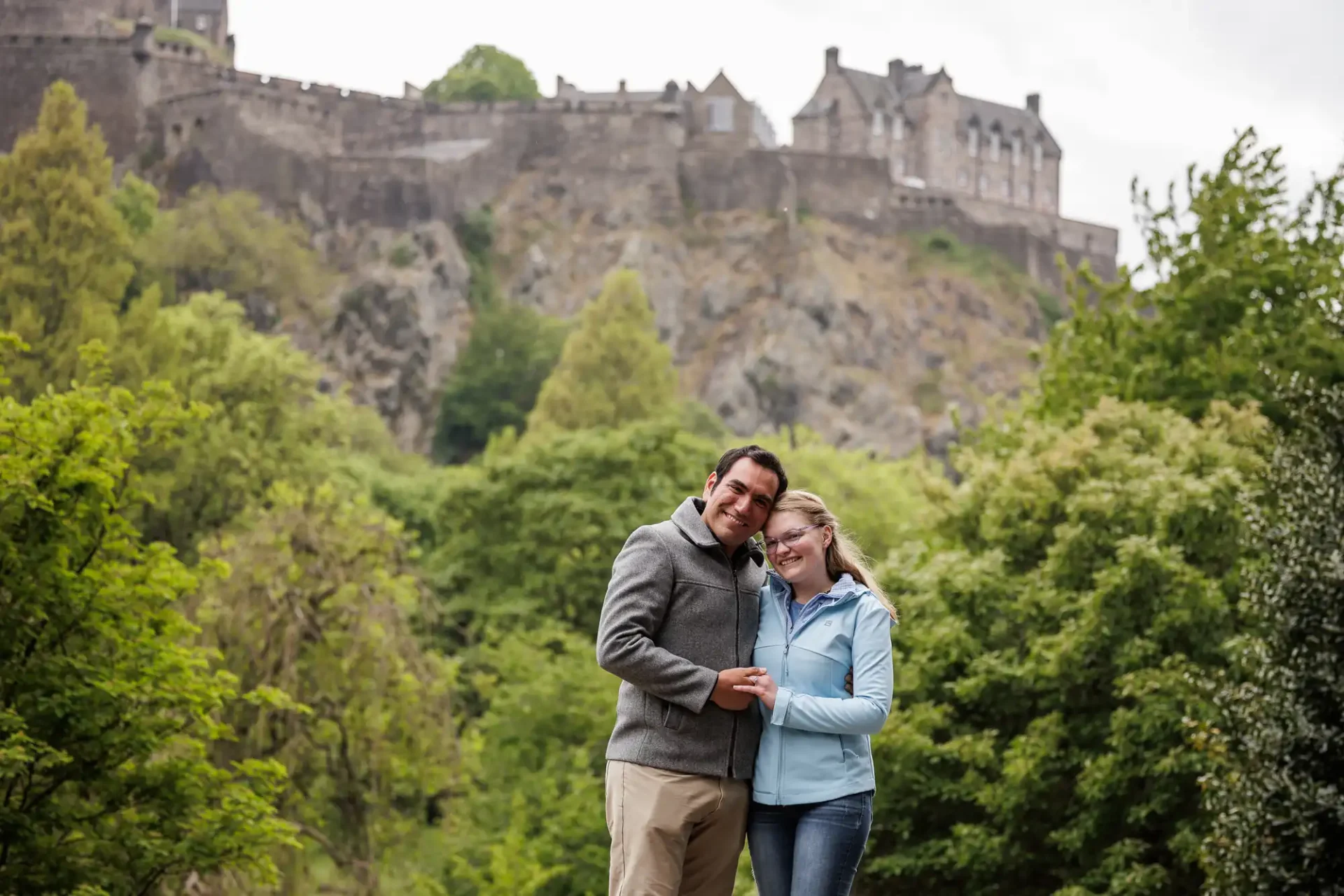A man and a woman stand closely together, smiling, in front of lush greenery with a large stone castle visible on a hill in the background.
