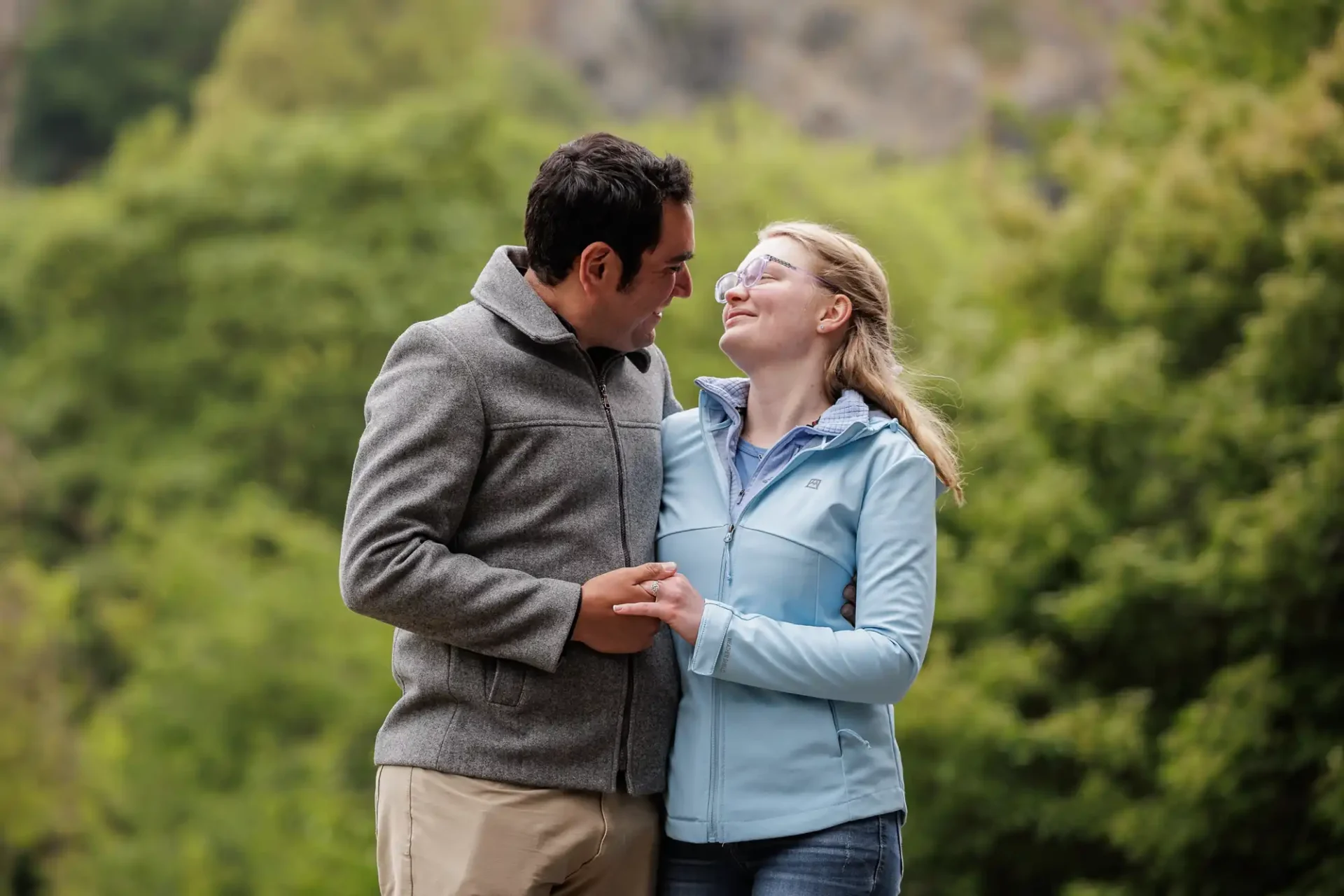 A man and woman stand outdoors, looking at each other and holding hands, with trees and greenery in the background.