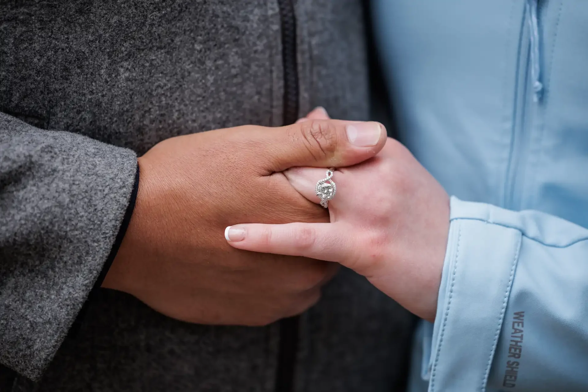 Two people hold hands, one wearing a grey coat and the other a light blue jacket, with a diamond engagement ring visible on one person's finger.