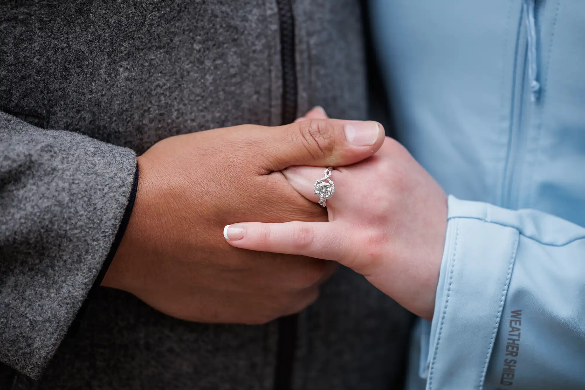 Two people hold hands, one wearing a grey coat and the other a light blue jacket, with a diamond engagement ring visible on one person's finger.