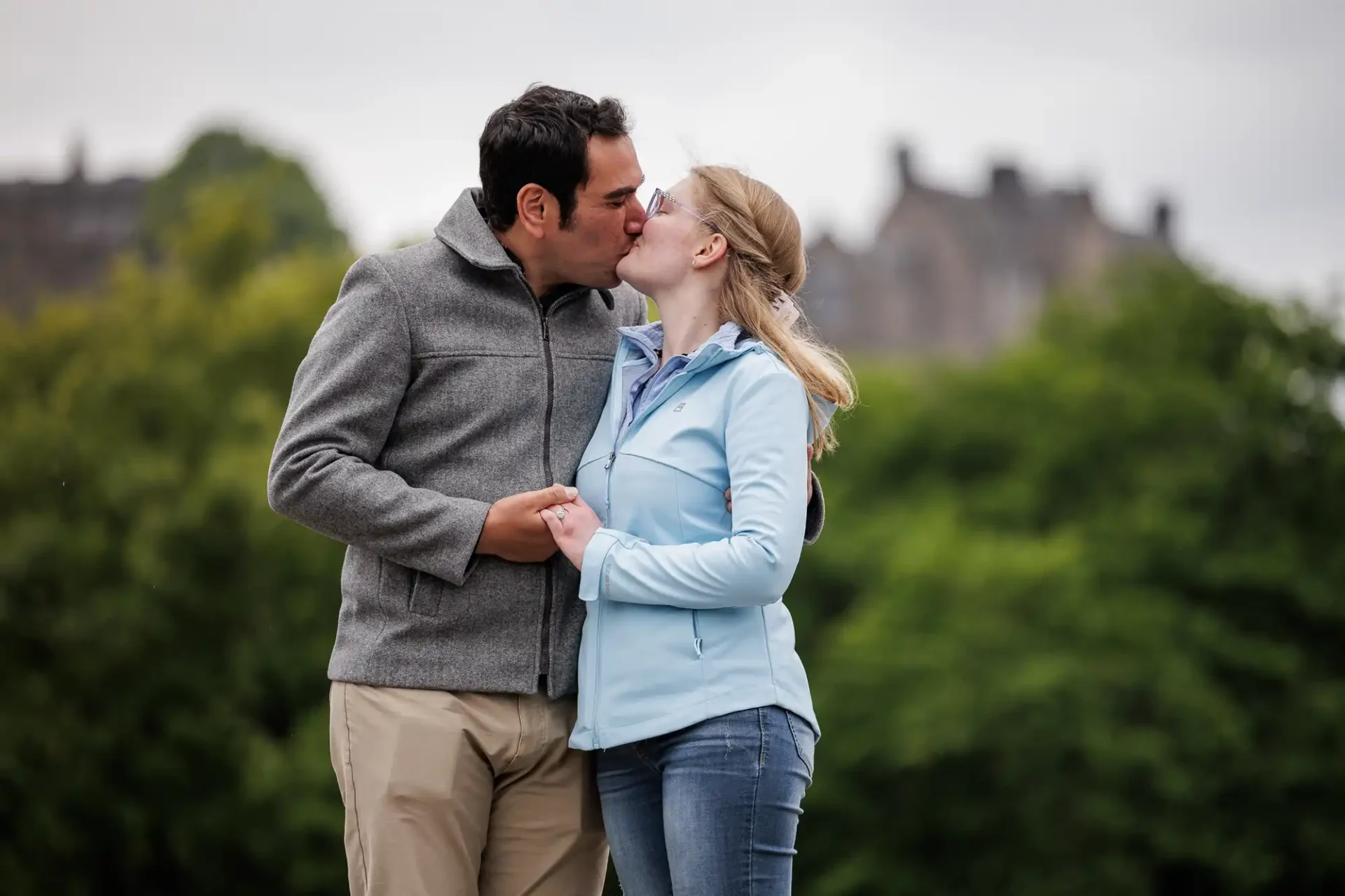 A man and woman stand outdoors, holding hands and kissing, with greenery and blurred buildings in the background.