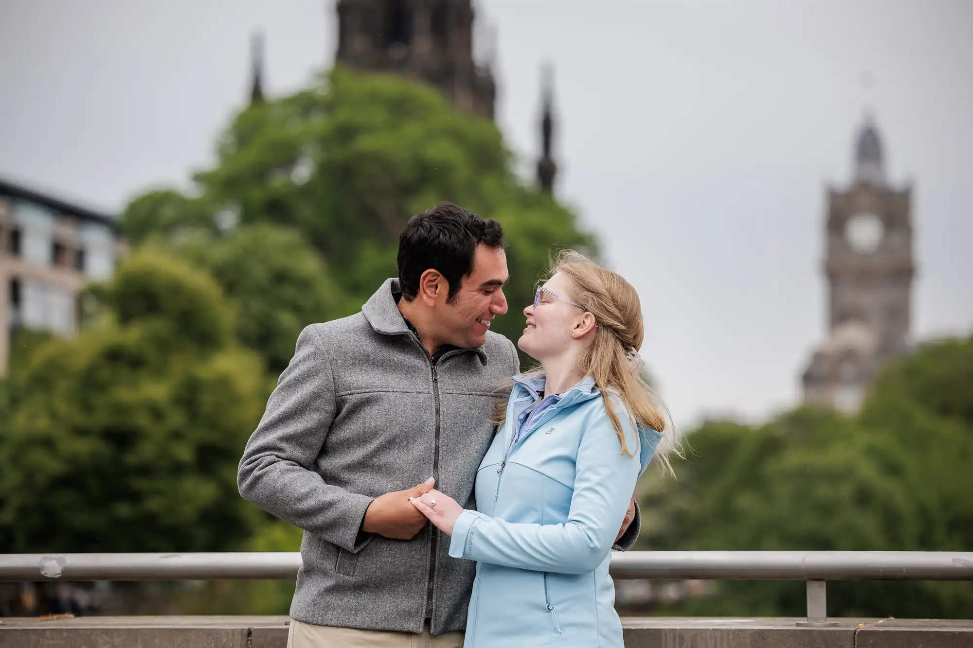 A man and a woman stand close together on a bridge, smiling at each other, with trees and historic buildings in the background.