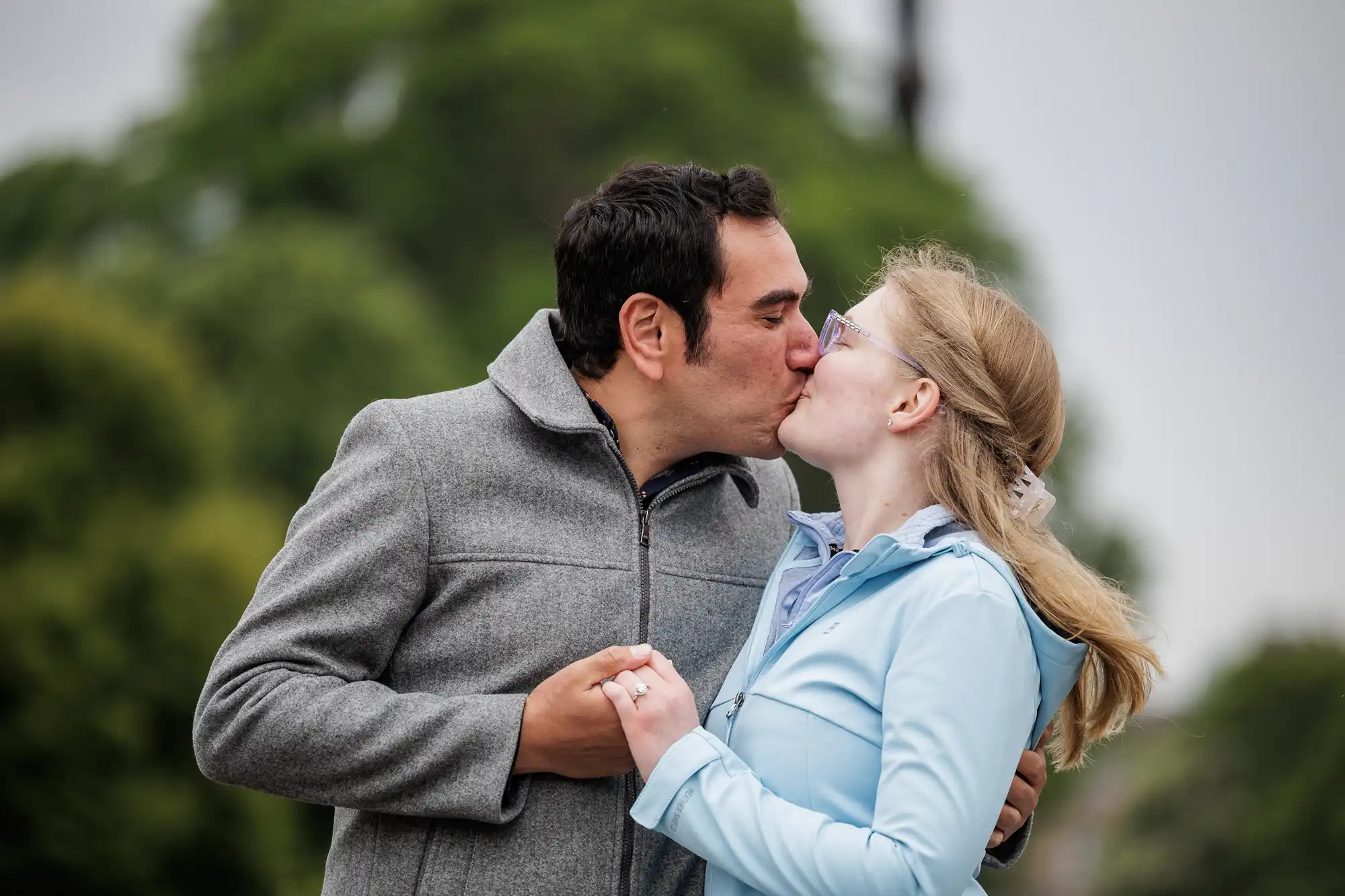 A man and a woman kiss outdoors while holding hands, with trees and a cloudy sky in the background.