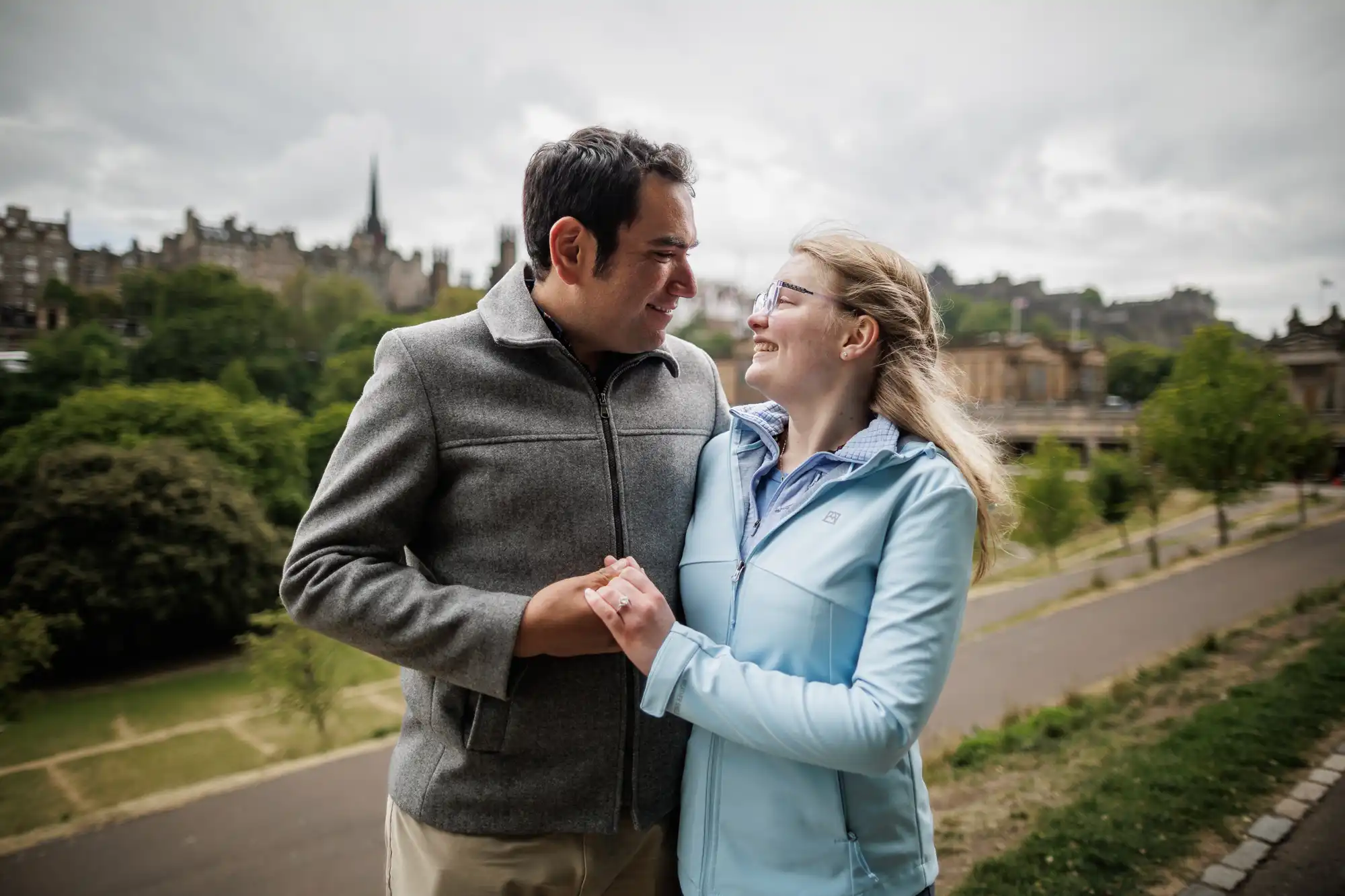 A man and a woman stand outdoors holding hands and smiling at each other, with historic buildings and greenery in the background.