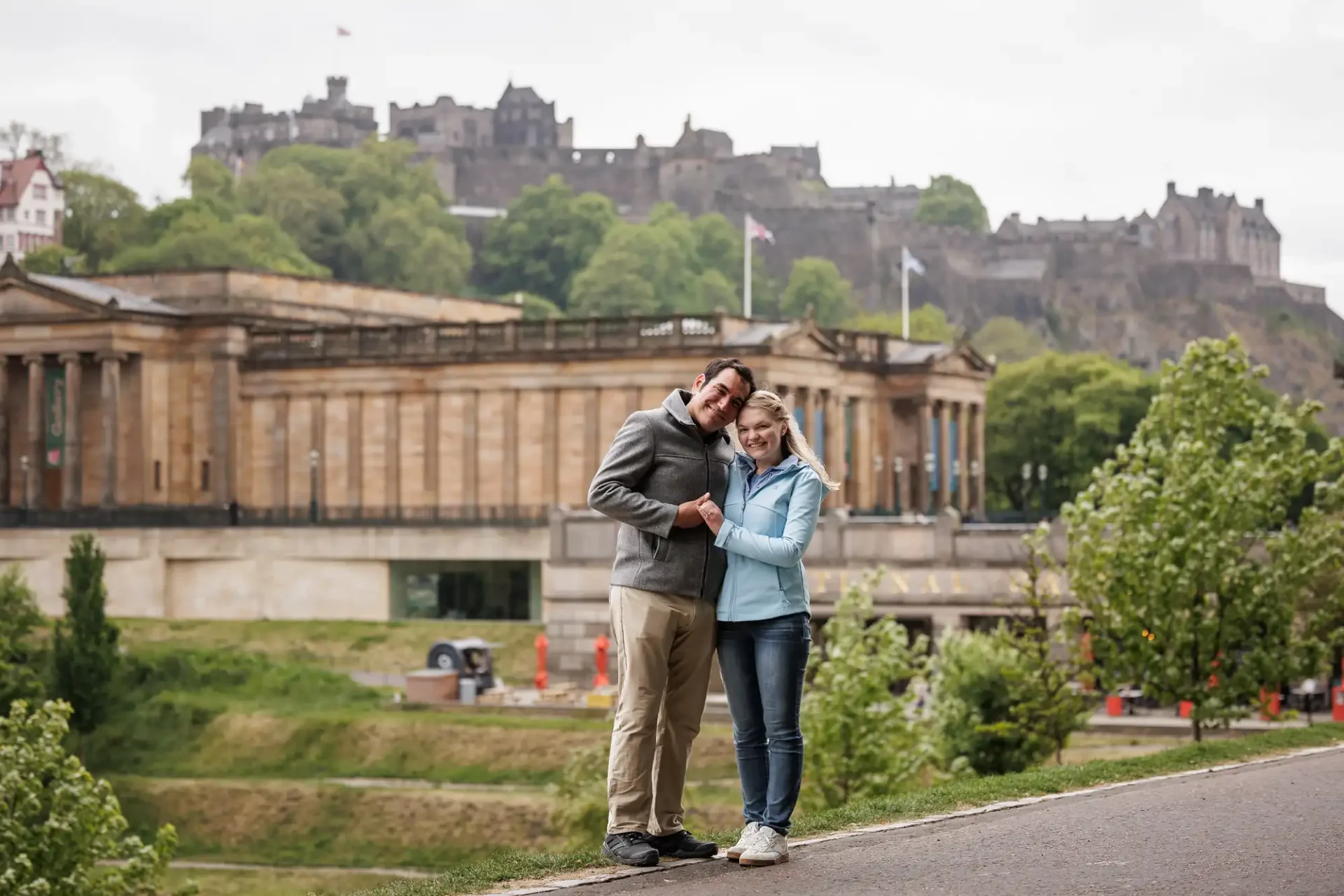 A smiling couple stands close together on a paved path with historic buildings and a castle on a hill in the background.