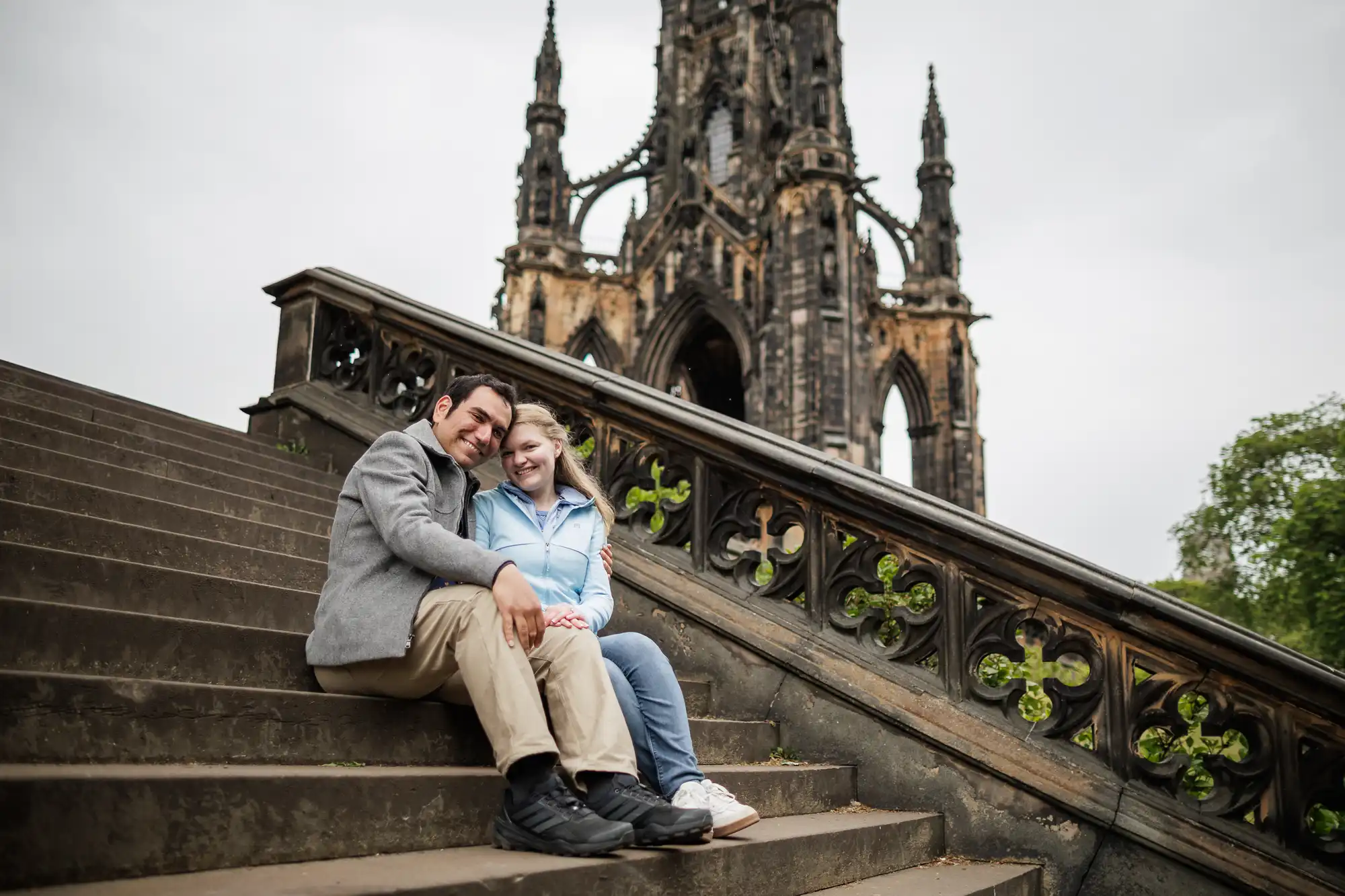 A couple sits together on stone steps in front of a large Gothic monument on a cloudy day.