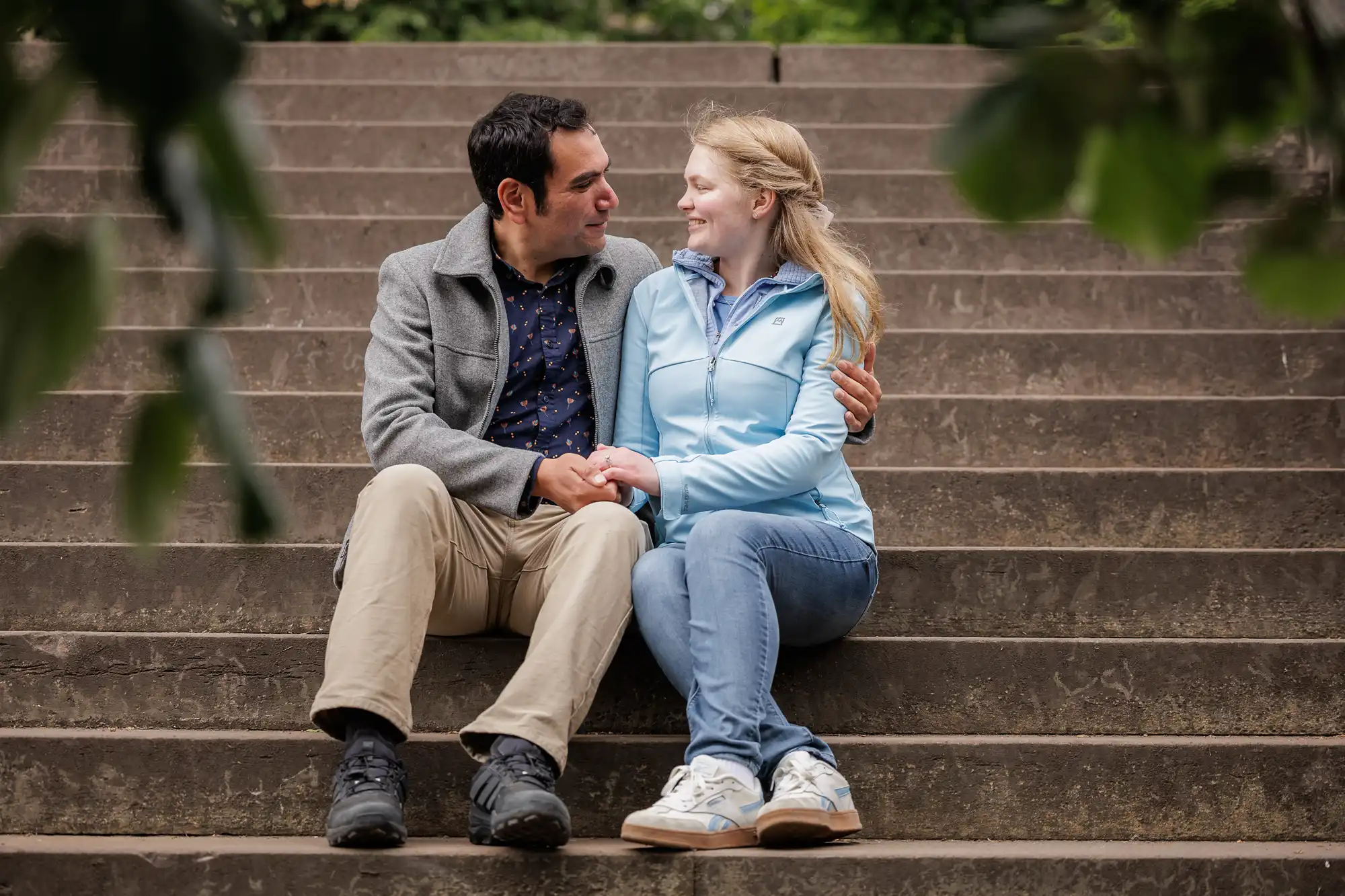 A man and a woman sit on outdoor steps, holding hands and looking at each other, surrounded by greenery.