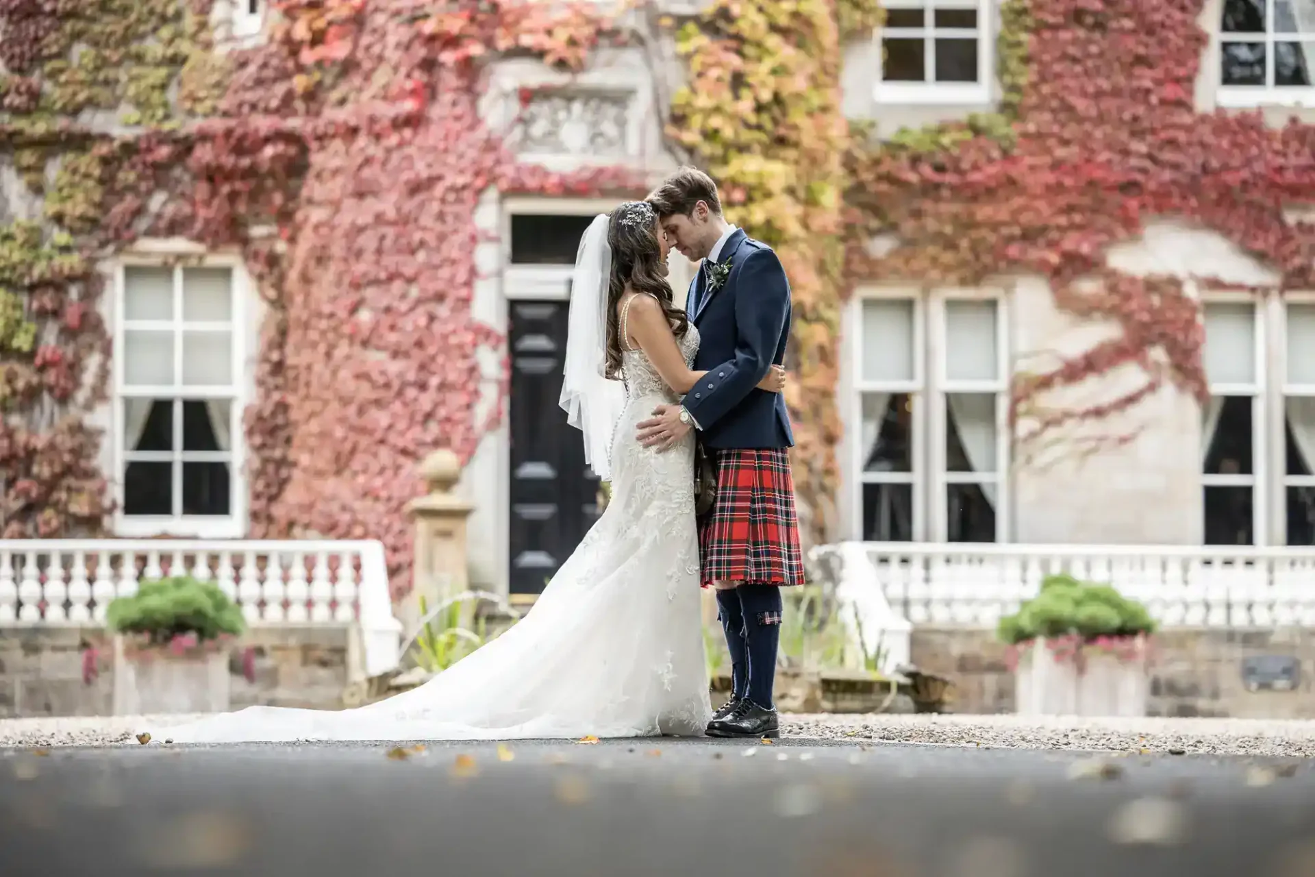A bride and groom stand close together outside a vine-covered building, with the groom wearing a kilt and the bride in a white wedding dress and veil at Carlowrie Castle, Edinburgh.