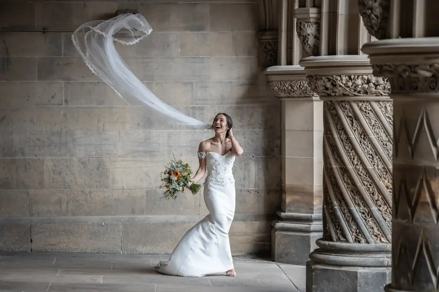 A bride in a white dress stands indoors, holding a bouquet, as her veil floats in the air behind her near ornate stone columns at Fettes College, Edinburgh.
