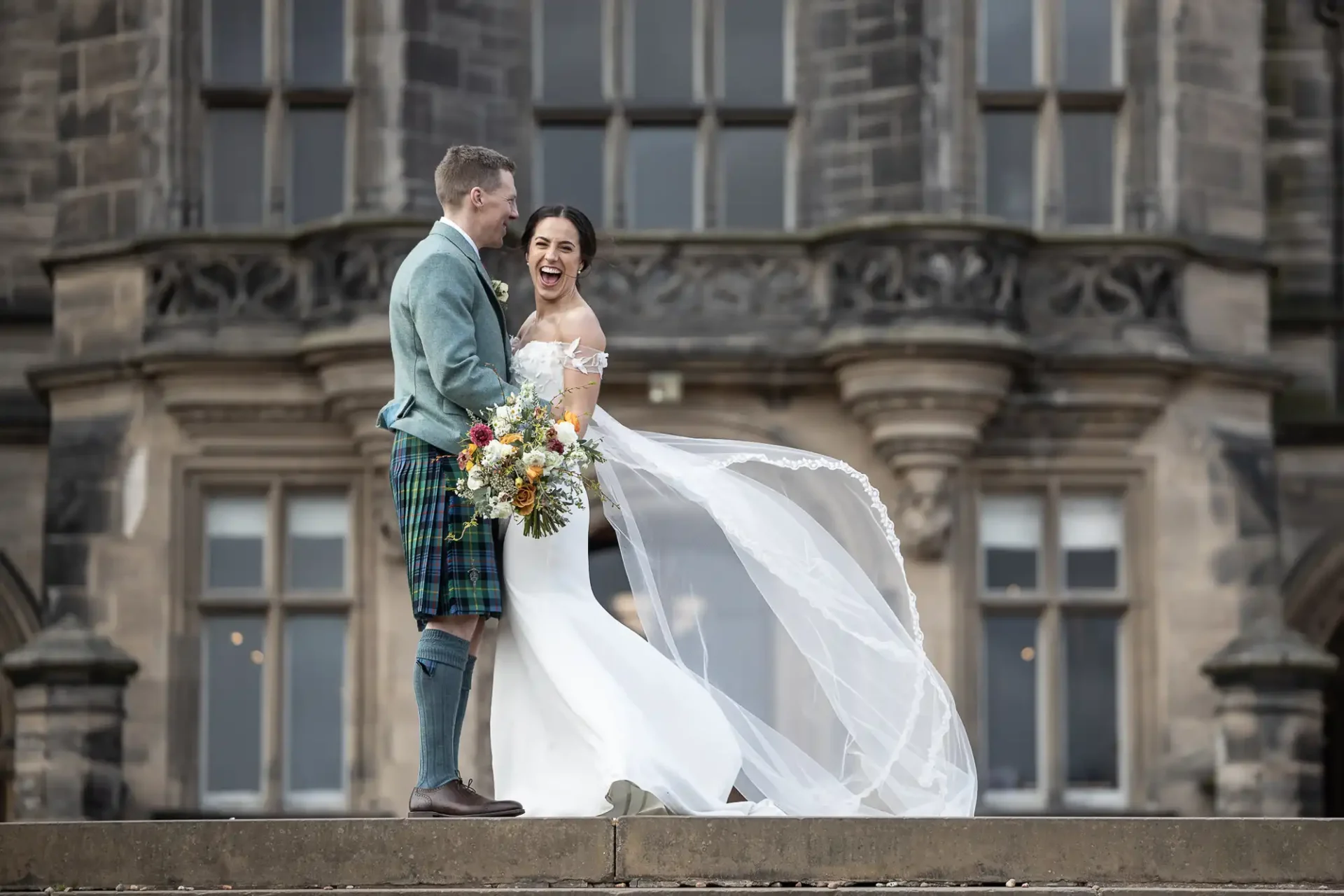 A bride in a white dress and veil stands smiling next to a groom in a kilt, holding a bouquet, in front of a stone building at Fettes College, Edinburgh.