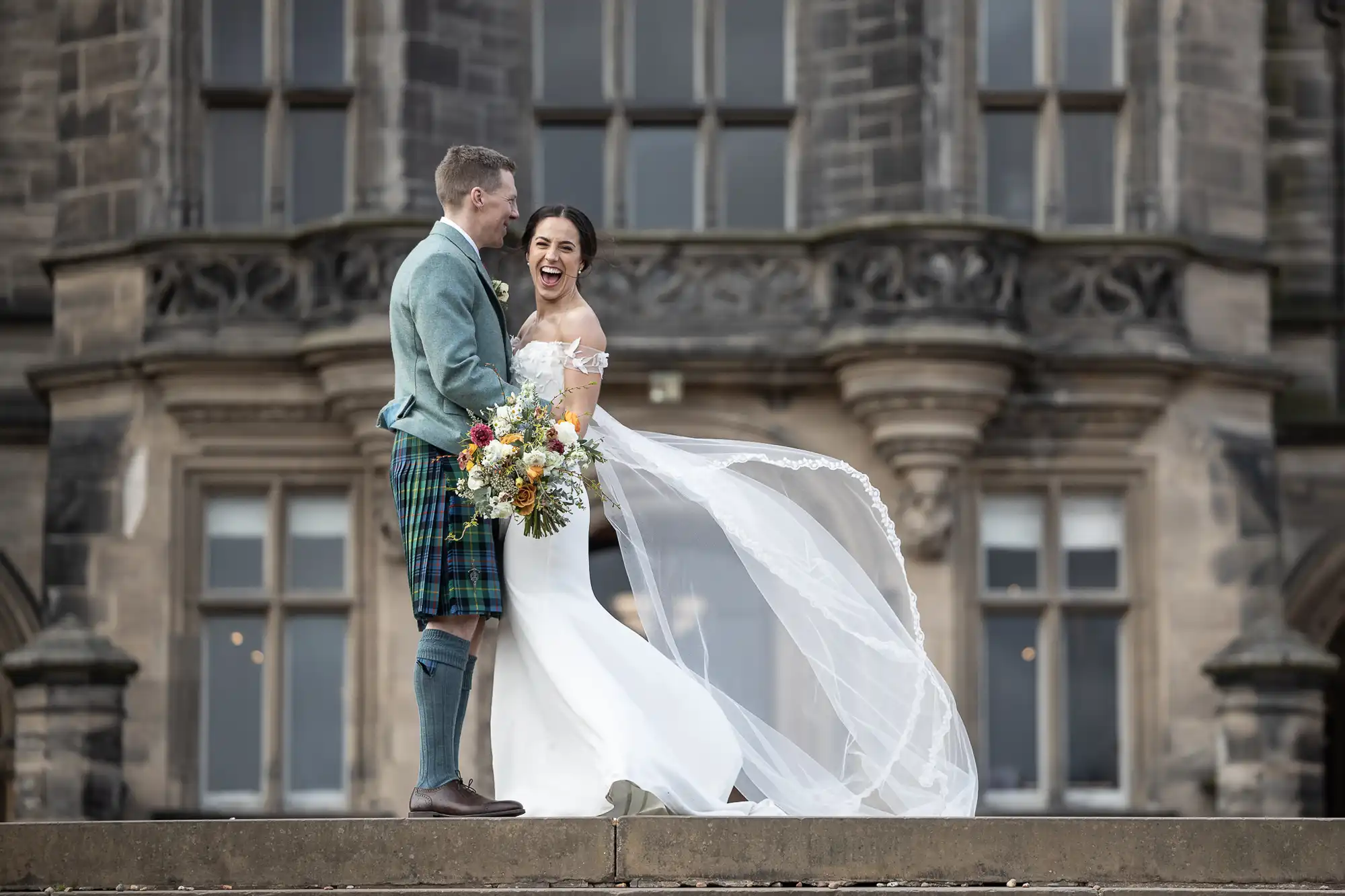 A bride in a white dress and veil stands smiling next to a groom in a kilt, holding a bouquet, in front of a stone building at Fettes College, Edinburgh.