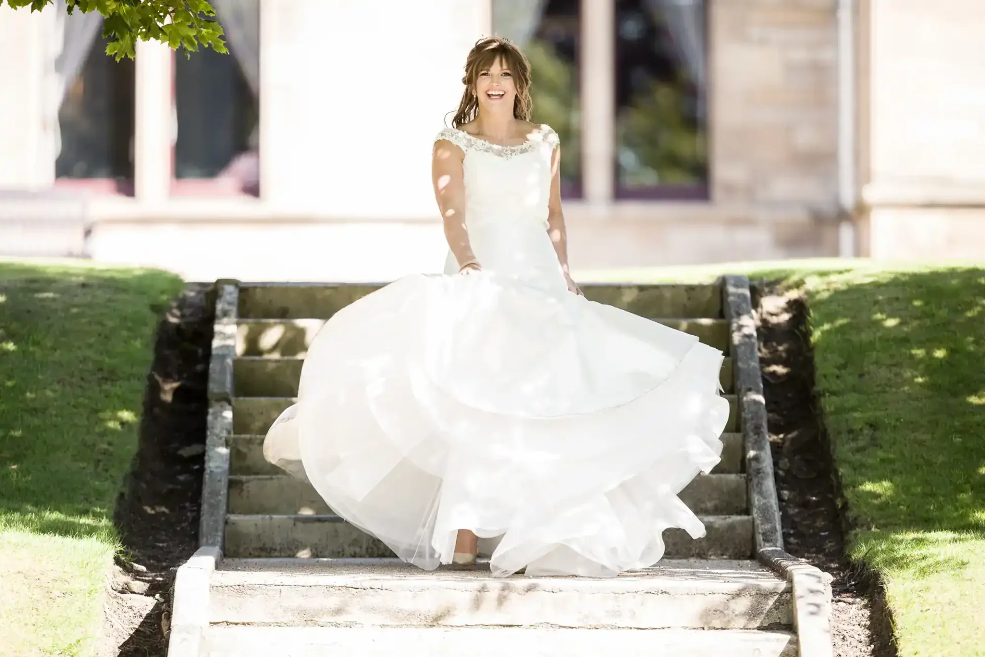 A woman in a white wedding dress stands on outdoor stone steps, holding her dress and smiling, with green grass and a building in the background at Napier University, Edinburgh.