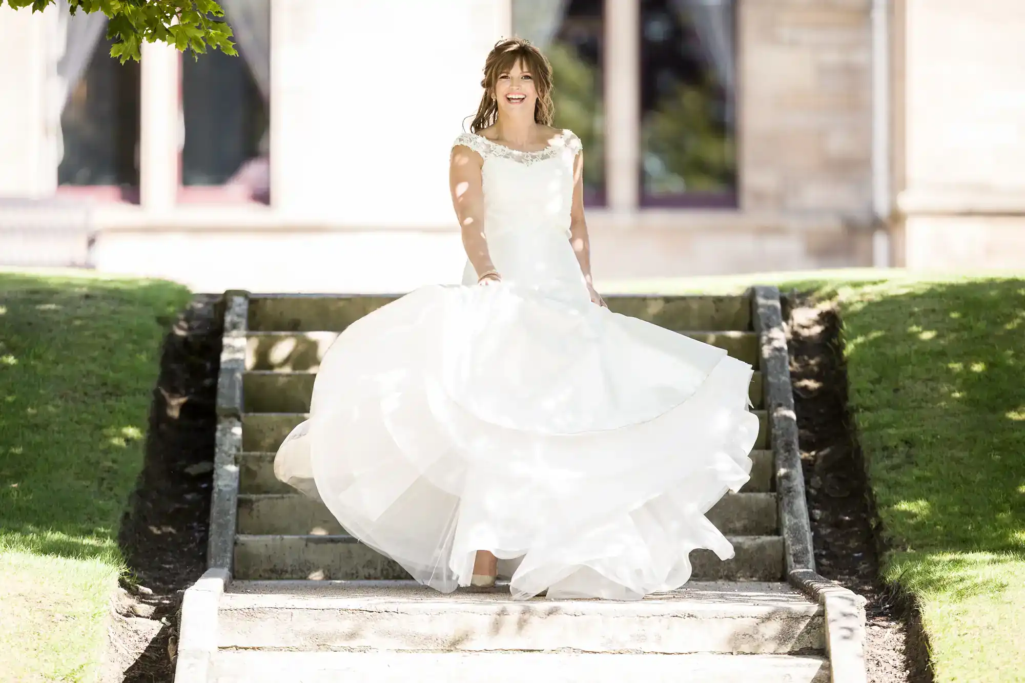 A woman in a white wedding dress stands on outdoor stone steps, holding her dress and smiling, with green grass and a building in the background at Napier University, Edinburgh.