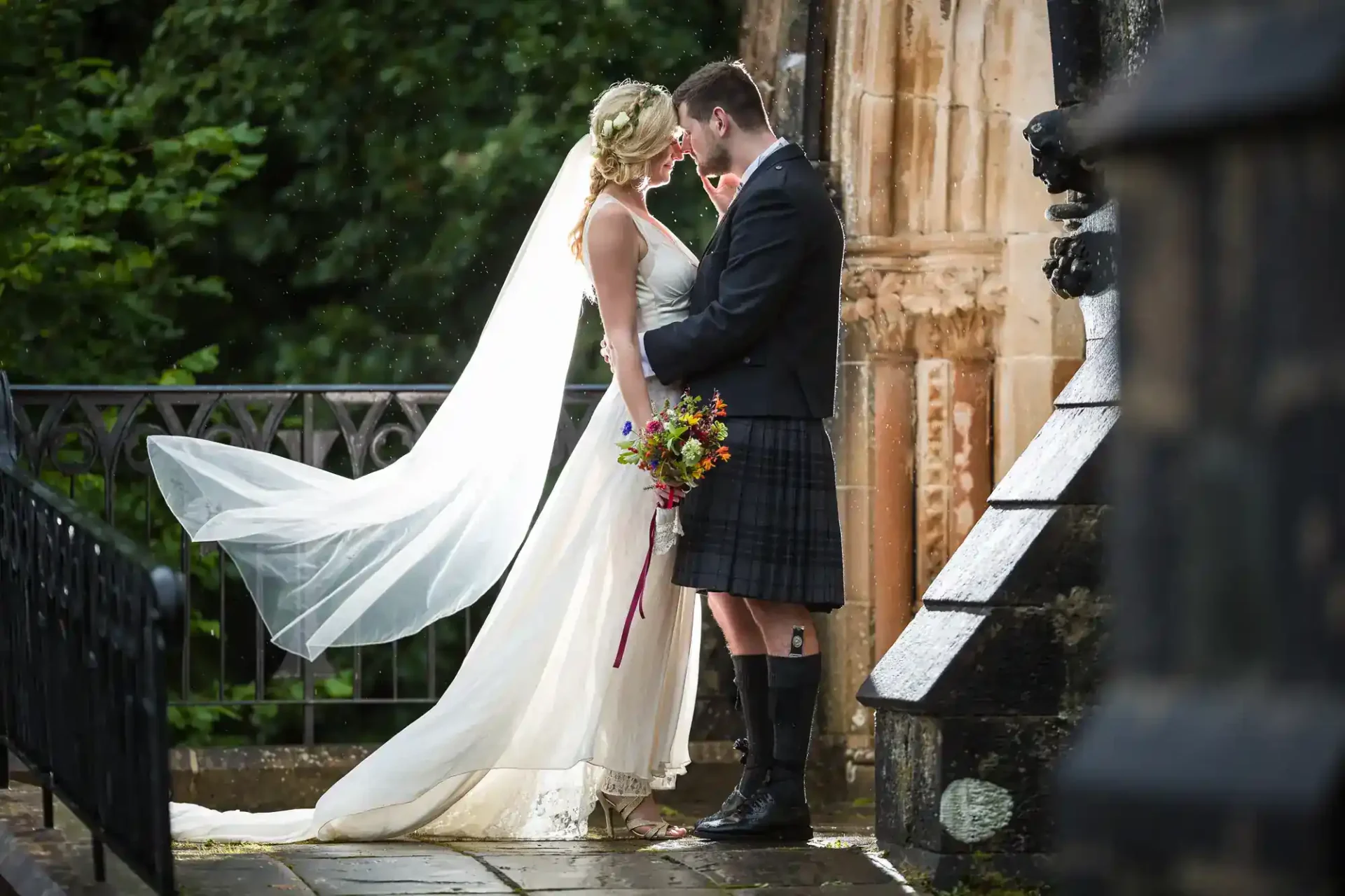 A bride and groom stand close together outside, with the bride holding a bouquet and her veil flowing behind her. The groom is wearing a traditional kilt at St John's Church, Edinburgh.