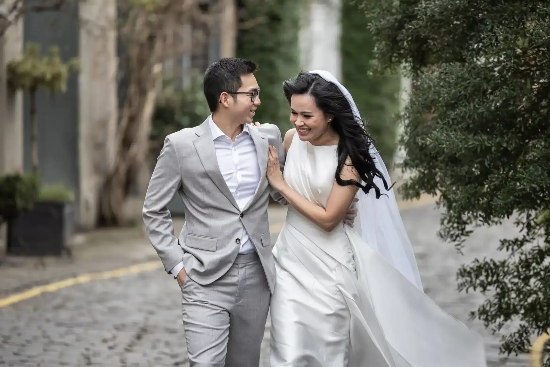 A bride in a white dress and veil walks arm-in-arm with a groom in a light grey suit on a cobblestone street at Circus Lane, Edinburgh.