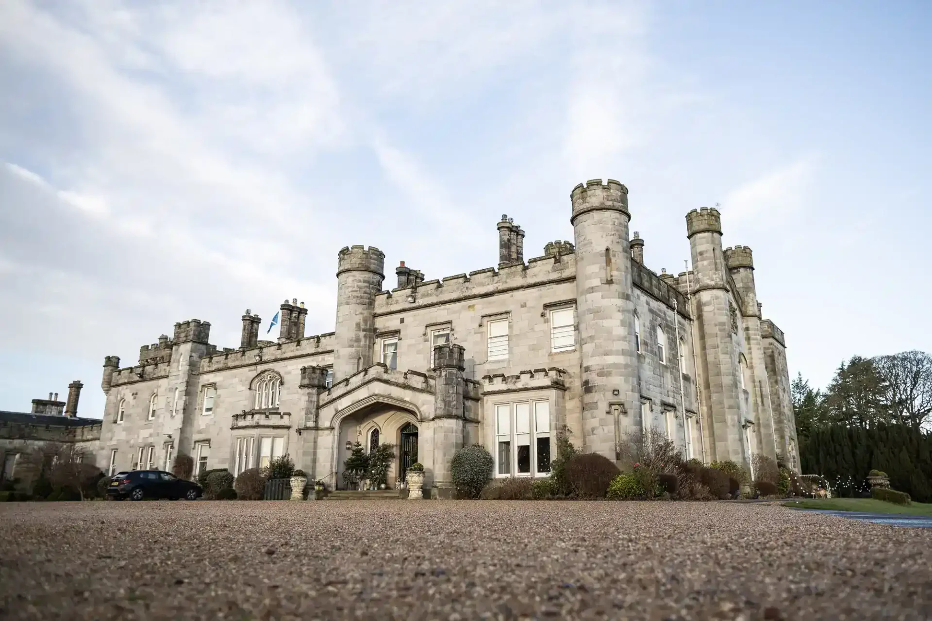 A large stone castle with turrets and arched entrance, surrounded by shrubs and a gravel driveway, under a partly cloudy sky at Dundas Castle, Edinburgh.