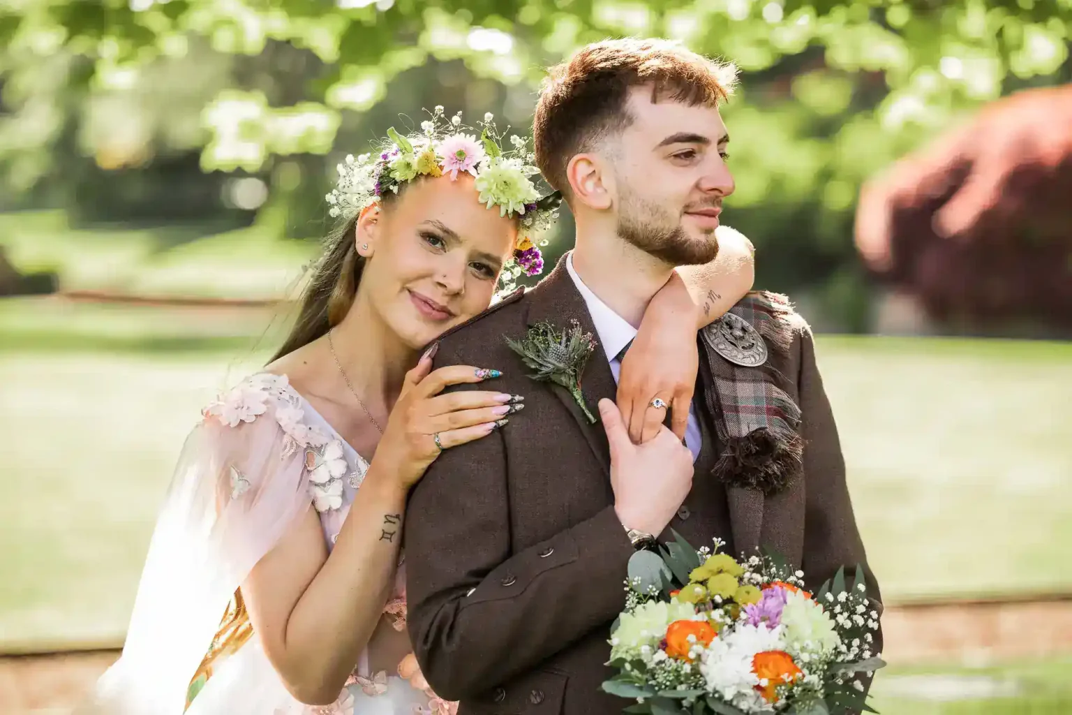 A woman in a floral dress and crown embraces a man in a suit holding a bouquet, both standing outdoors in a green, sunlit garden at Prestonfield House, Edinburgh.