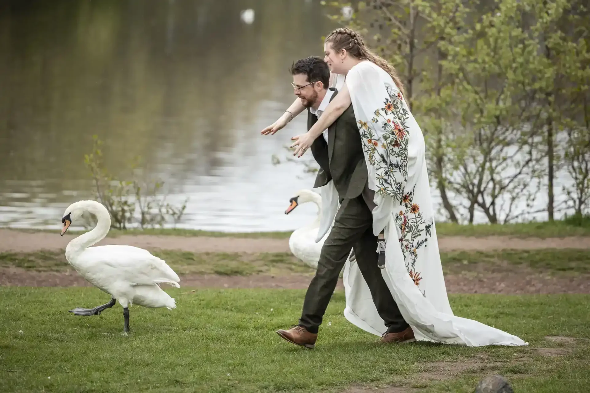 A bride in a floral gown rides piggyback on a man in a suit, reaching toward two swans by a lake in a park at St Margaret's Loch, Edinburgh.