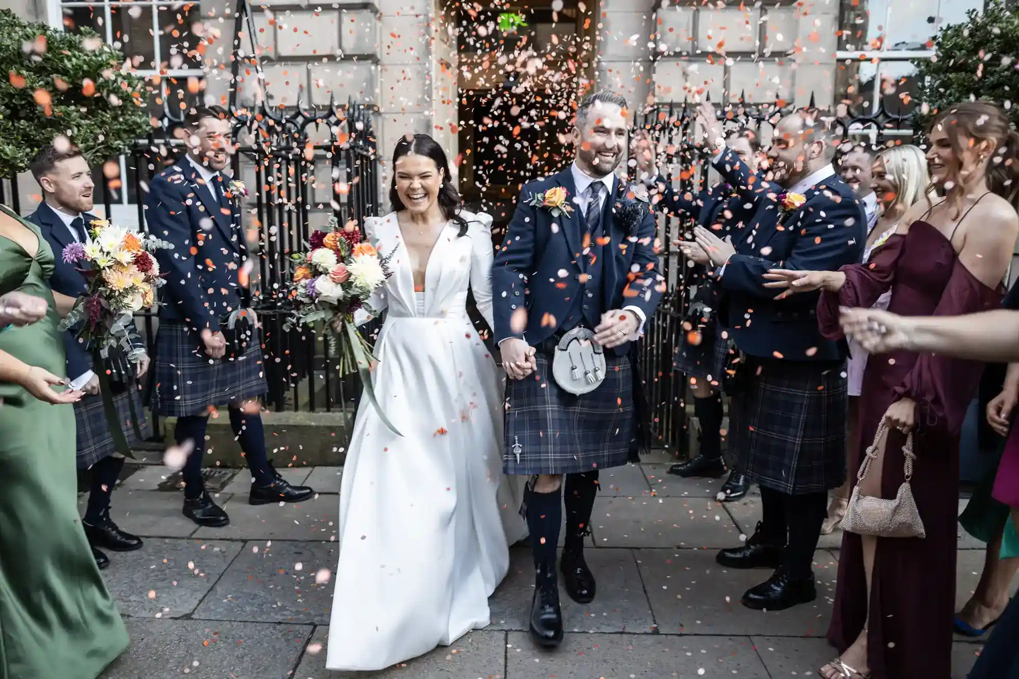 Wedding Content Creator image of a smiling bride in a white gown and groom in a kilt walk hand-in-hand as guests throw flower petals. The groom and wedding party wear kilts and suits, and many guests hold bouquets. They celebrate outside a stone building with black railings, the Signet Library.
