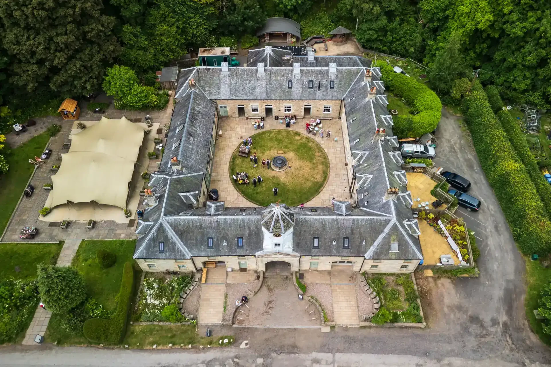 Aerial view Falkland Estate wedding reception featuring of a large, square stone building with a central grassy courtyard and fountain, surrounded by forest. People are gathered in the courtyard. Outdoor seating with canopy is on the left side; parked cars and pathways are around the building.