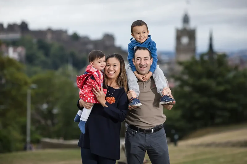 A couple stands outdoors with a young child in the woman’s arms and an older child on the man’s shoulders. A townscape is visible in the background.