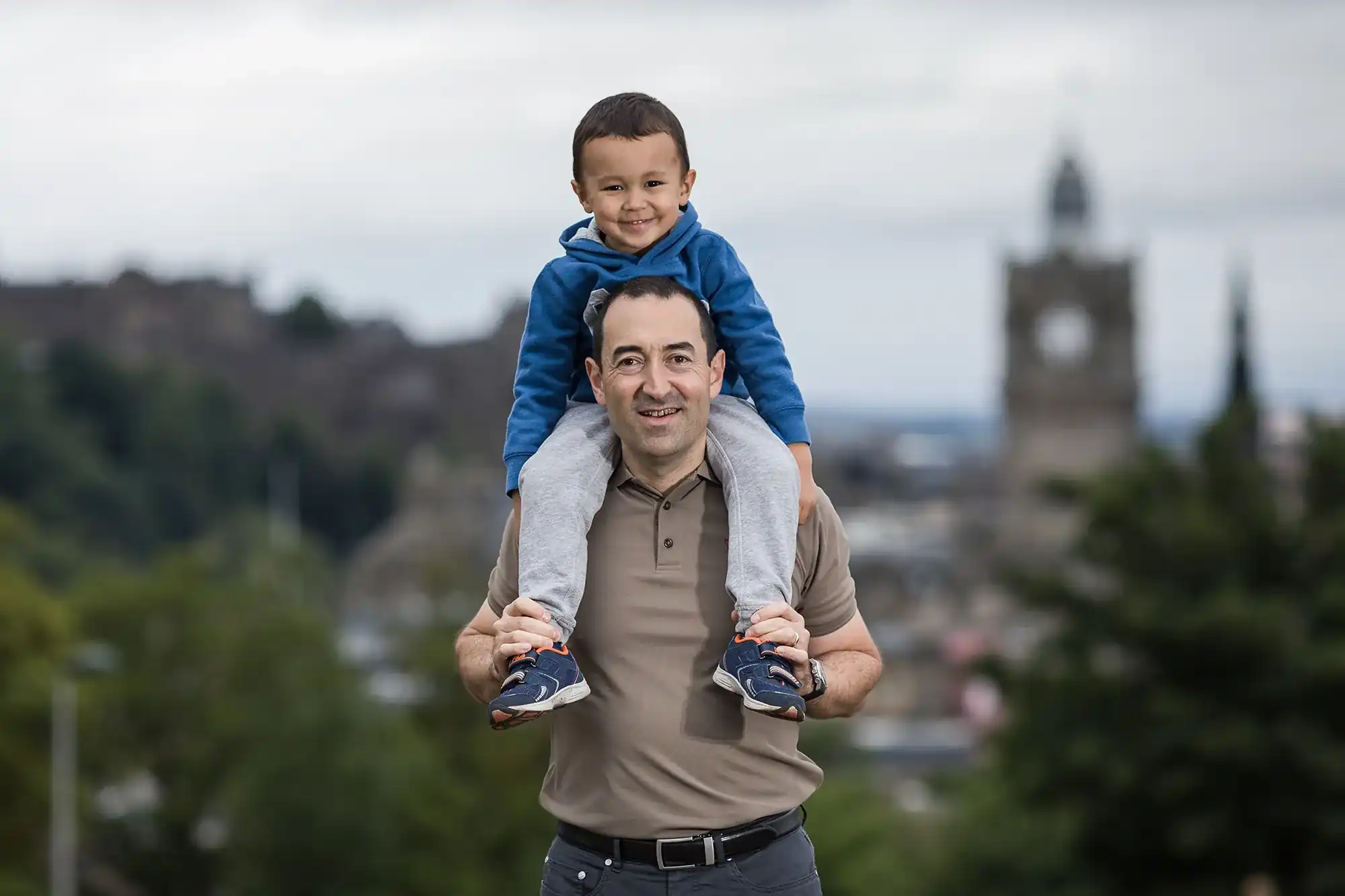 A man carries a smiling child on his shoulders outdoors. A blurred building and greenery are visible in the background.