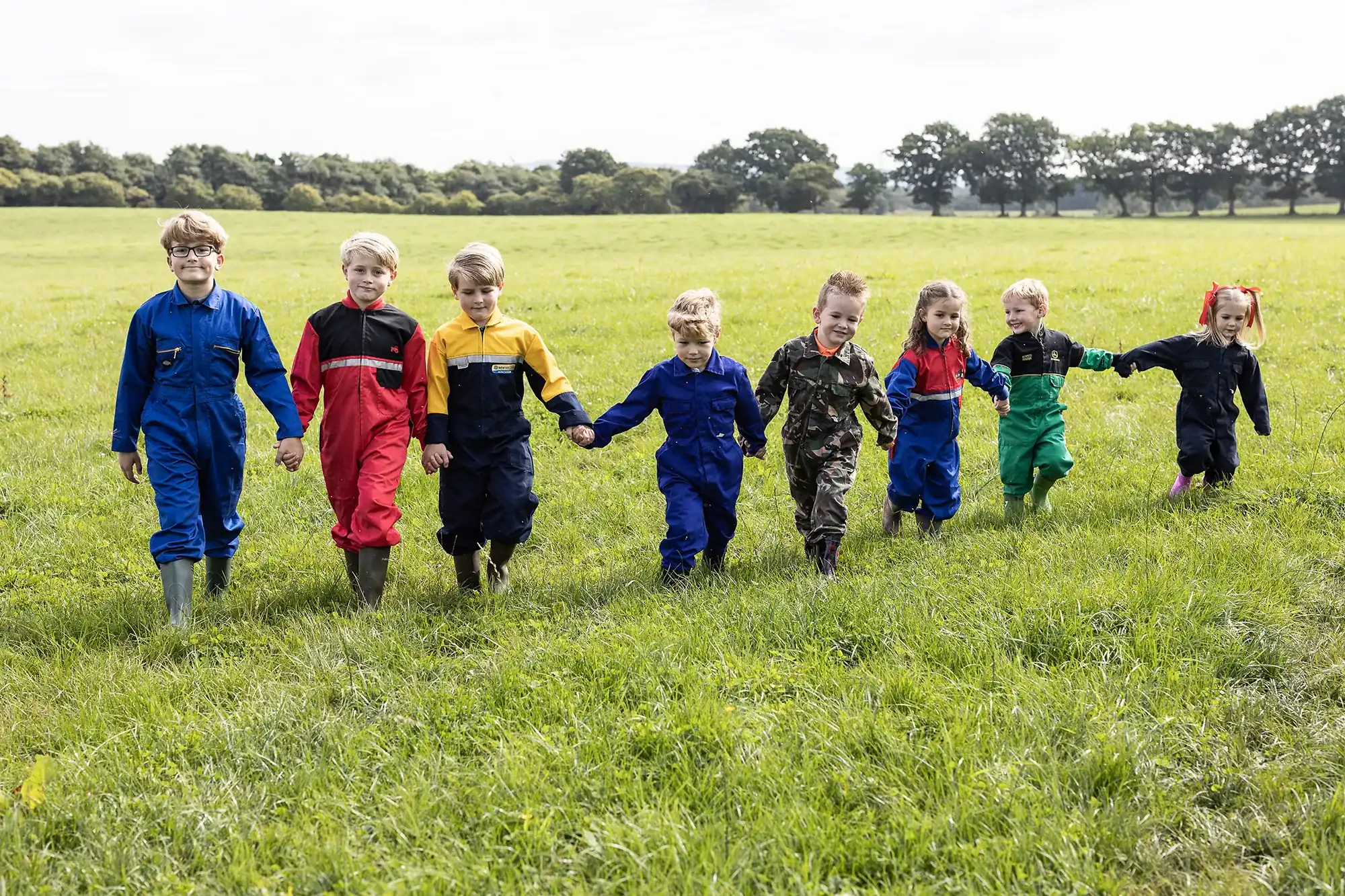 A group of children in colorful overalls and boots hold hands while walking in a grassy field with trees in the background.