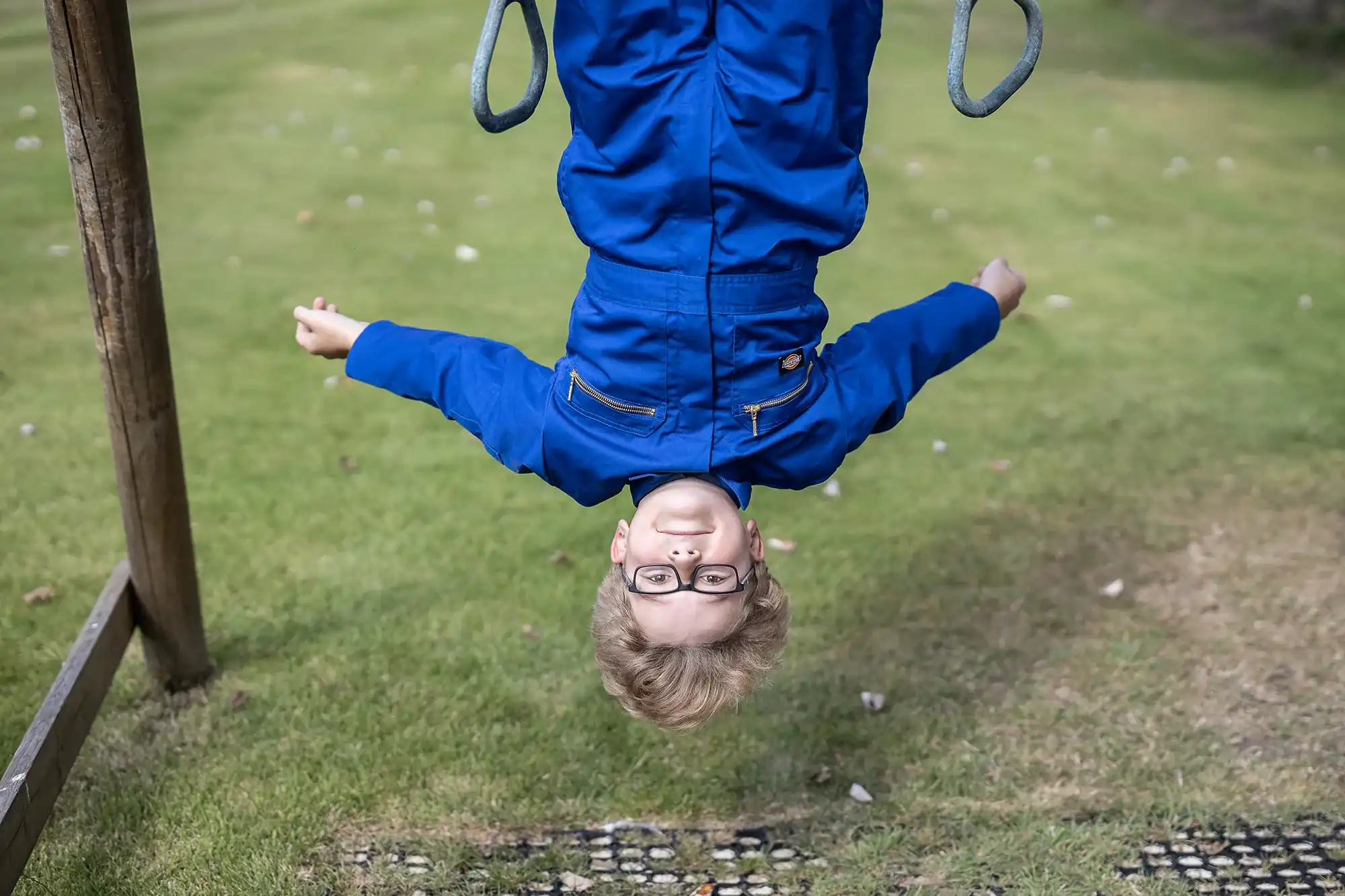 Child wearing glasses and a blue jacket hangs upside down from playground equipment with a grassy background.