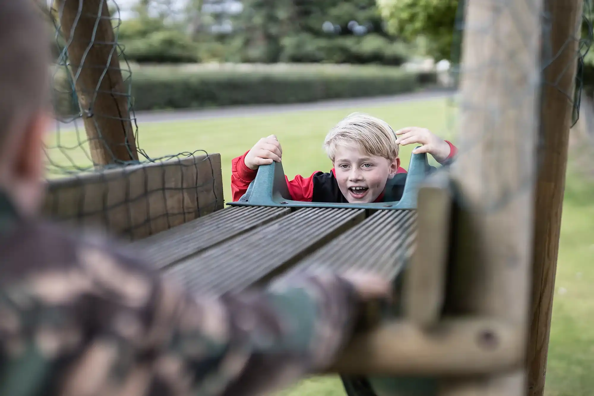 A child in a red jacket looks through a gap in a wooden play structure, with another child in camouflage clothing in the foreground.