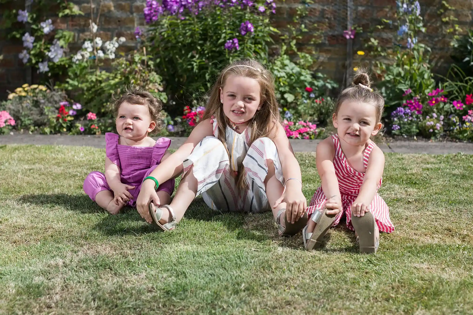 Three young children sit on grass in a garden with colorful flowers in the background, smiling at the camera. Two of them are wearing striped dresses, and one is in a purple outfit.