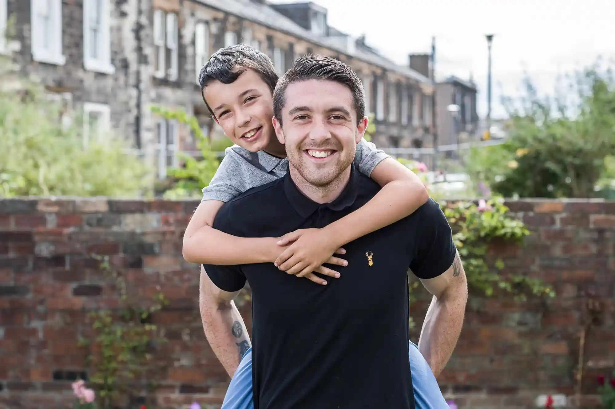 A man with short dark hair smiles as he gives a piggyback ride to a young boy with short hair. They are outdoors, with a brick wall and houses in the background.