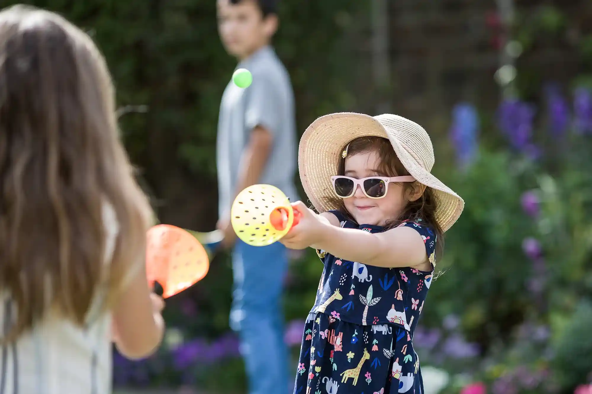 Two children playing in a garden. One child in a hat and sunglasses is holding a toy racket, while the other faces away. A third child stands in the background. Flowers and greenery are visible.