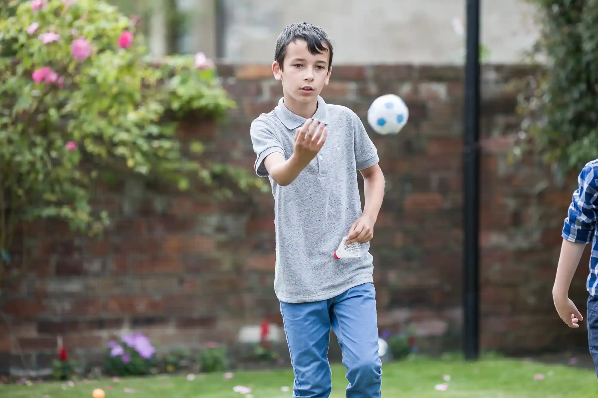 A boy in a gray polo shirt and blue pants catches a small soccer ball in a garden with green grass and a brick wall in the background.