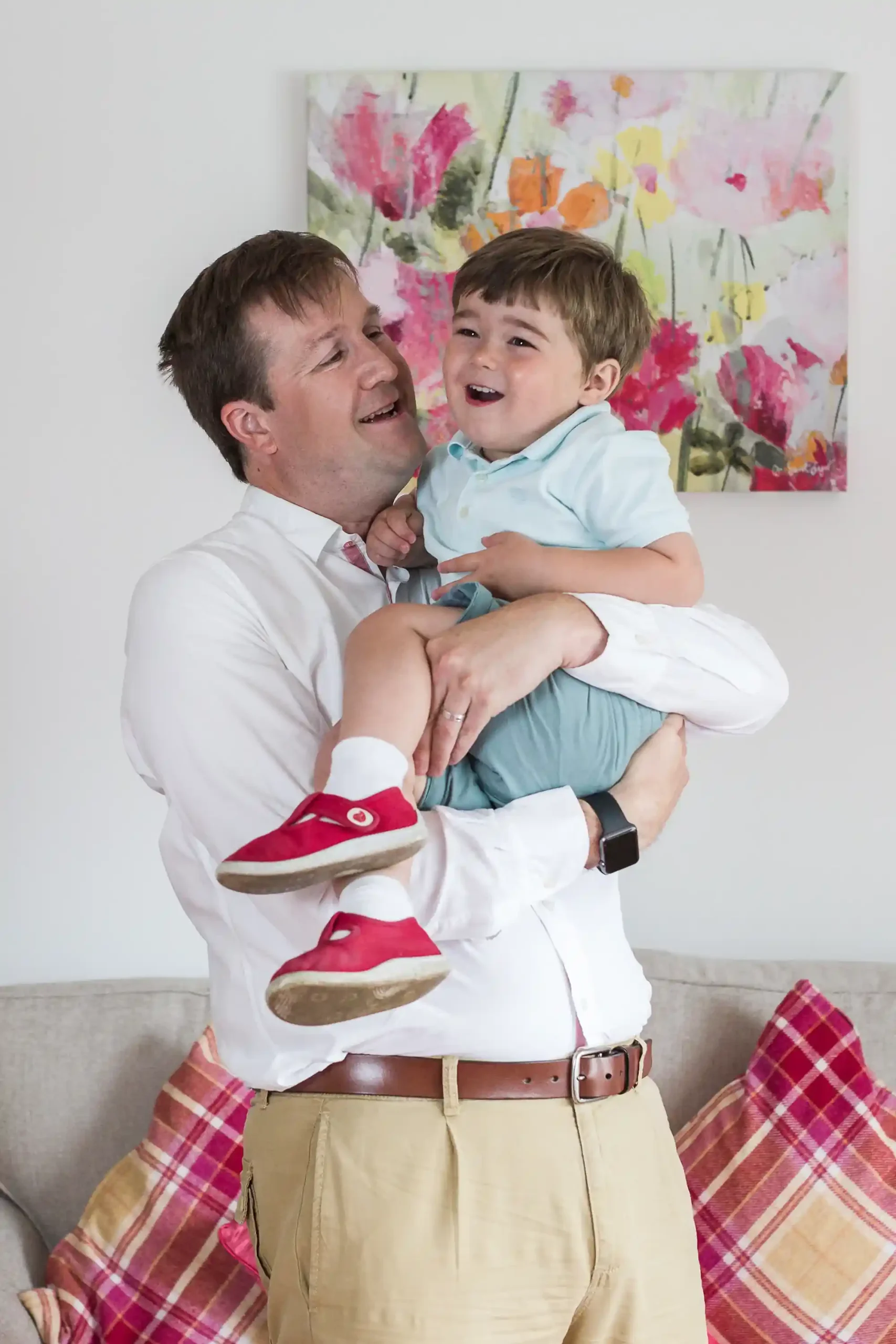 A man in a white shirt and beige pants is holding and smiling at a young boy in a blue shirt and red shoes. They are standing in a living room with a floral painting and plaid pillows.