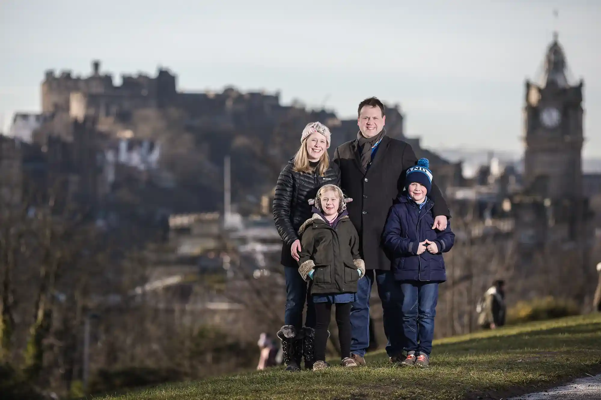A family of four poses outdoors with a historic castle and clock tower in the background.