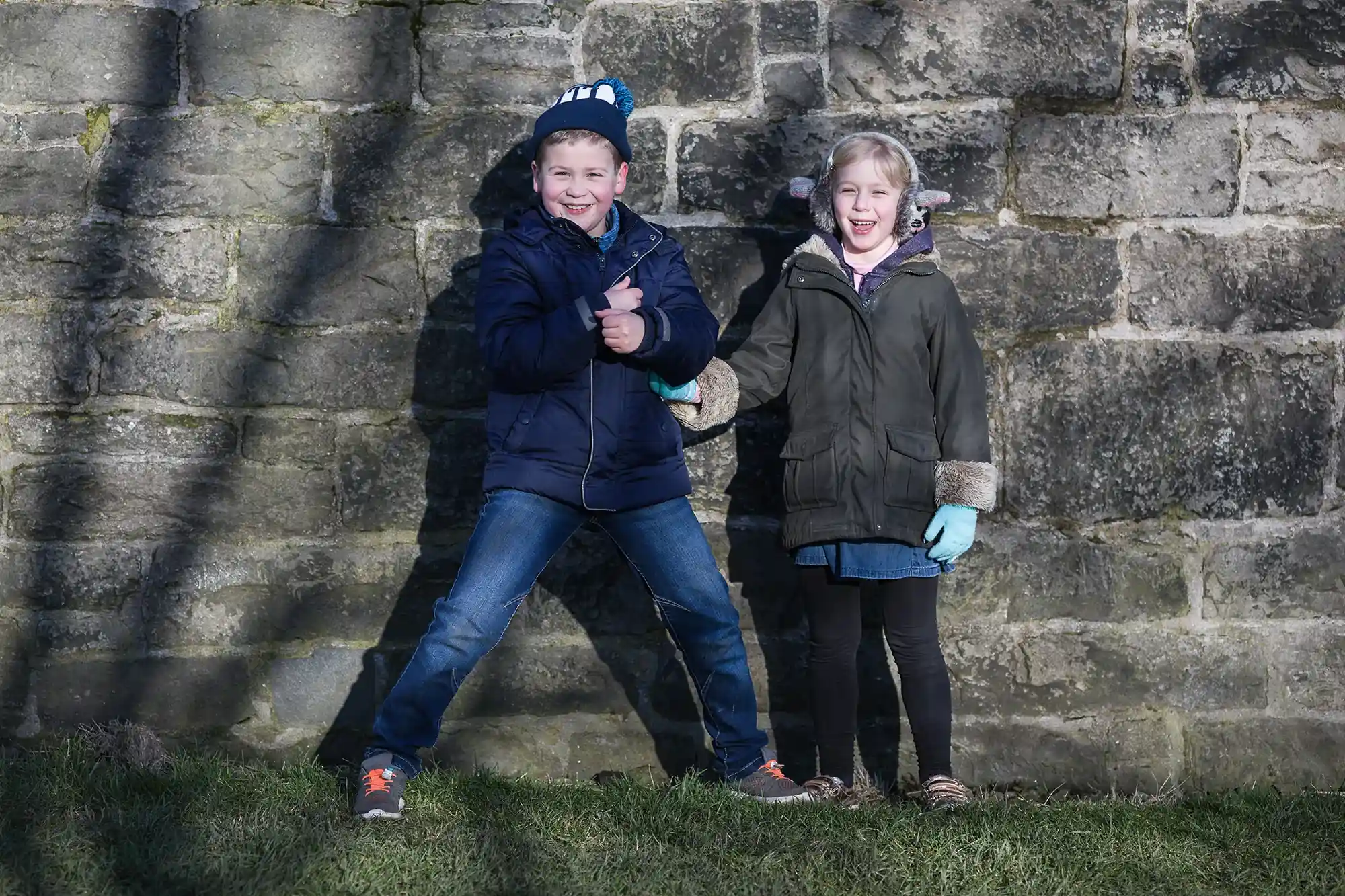 Two children stand in front of a stone wall, one giving a thumbs up and the other smiling. Both wear winter jackets and gloves, and shadows from a tree are cast on the wall behind them.