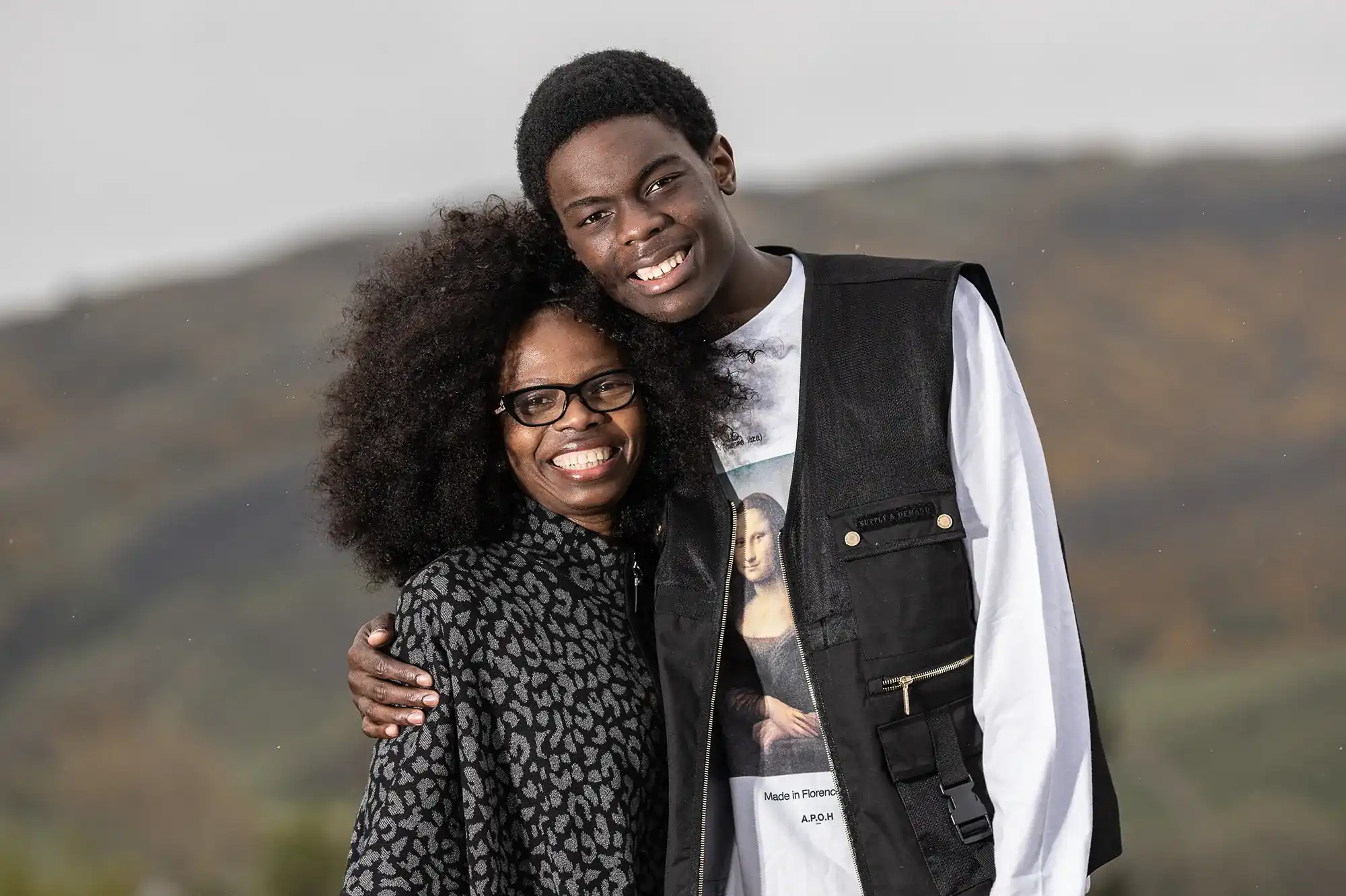 A smiling woman with curly hair and glasses is hugged by a taller smiling young man in front of a blurred outdoor background.