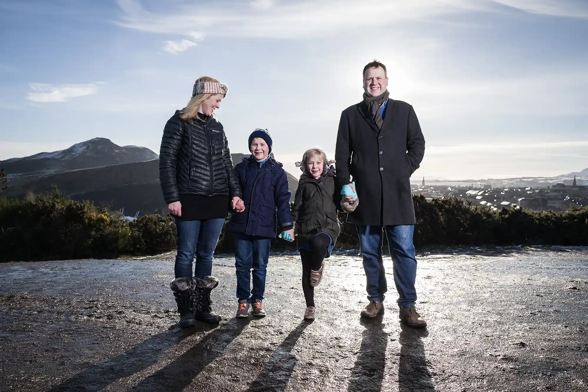 A family of four, two adults and two children, stand outdoors on a paved path with a scenic mountainous backdrop. The adults wear coats, and the children wear jackets and hats. One child lifts a leg playfully.