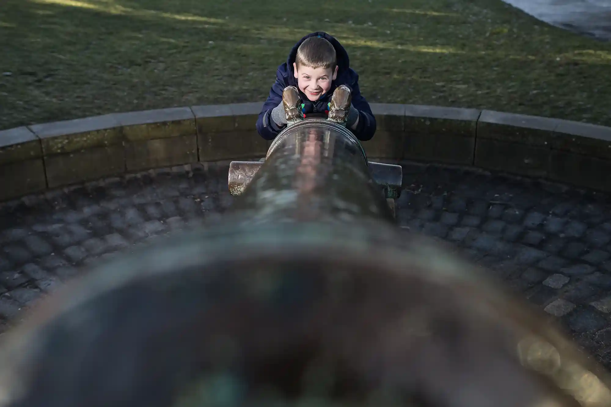 A child is smiling while looking through the barrel of an old cannon in an outdoor setting.