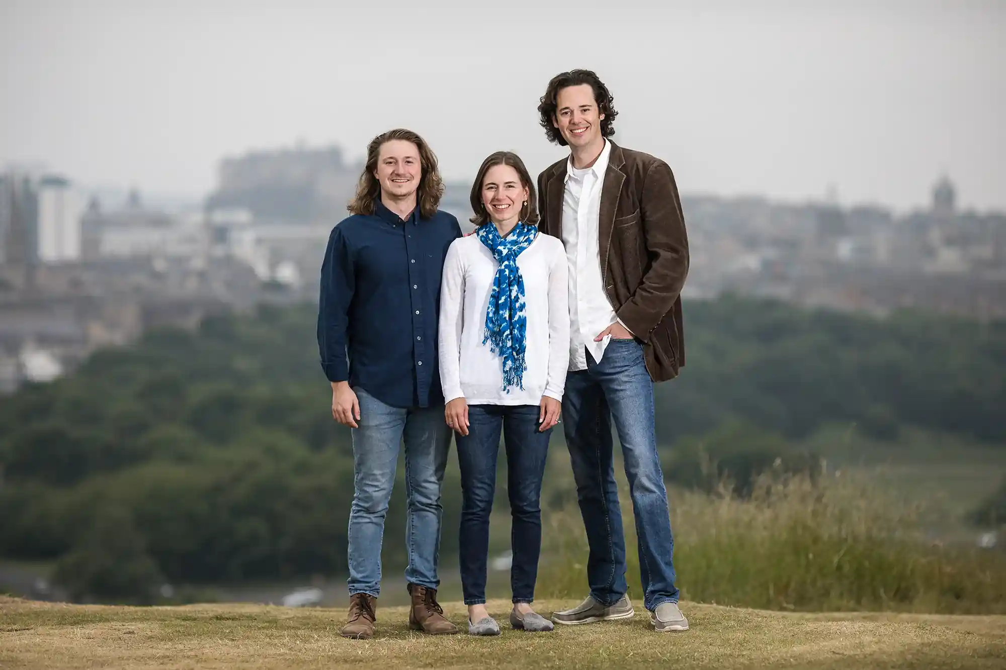 Three people stand outdoors on a hill with a cityscape in the background. They are casually dressed and smiling at the camera.
