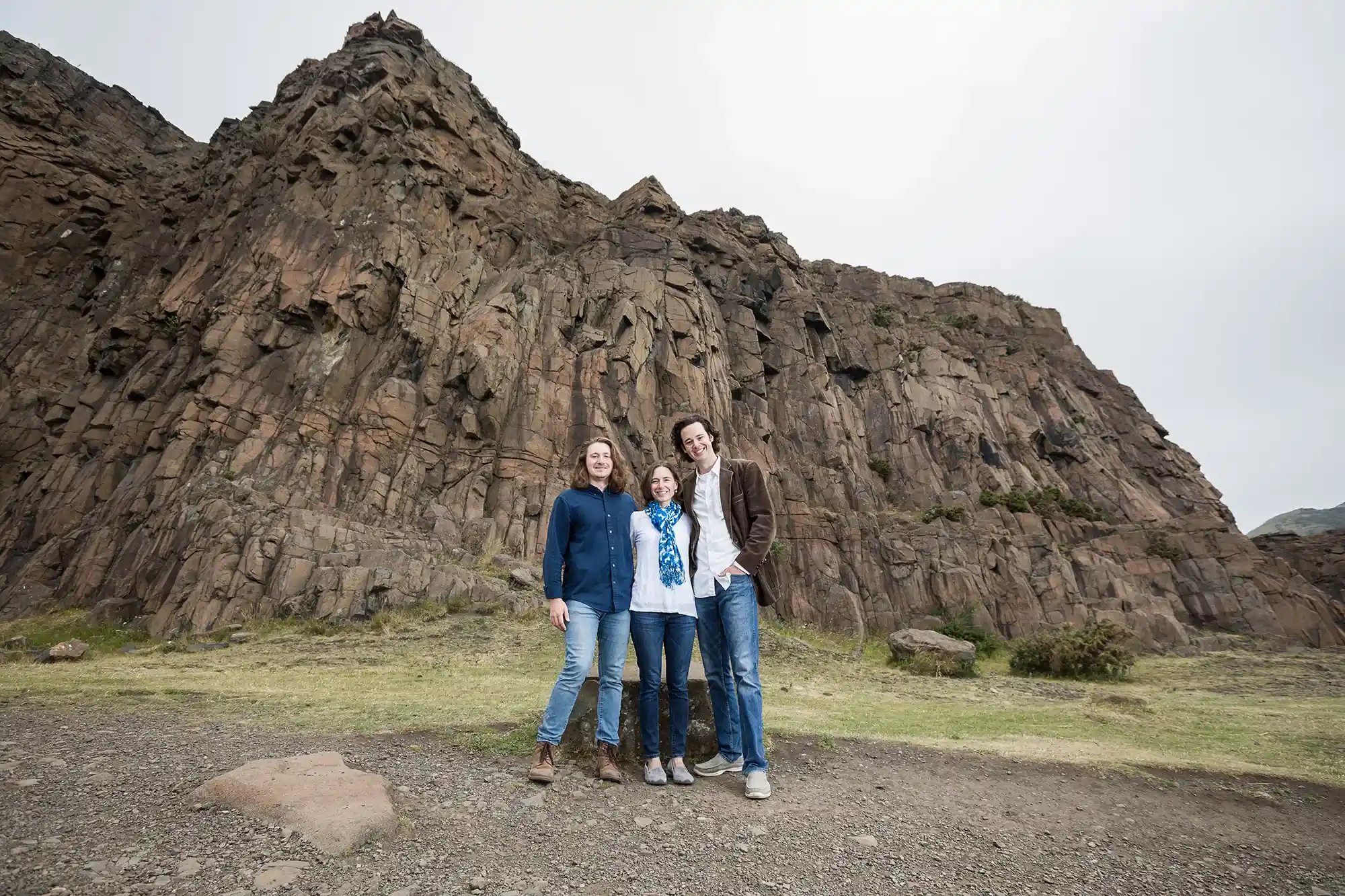 Three people standing together in front of a large rocky cliff face, with cloudy sky overhead. They are on a rocky and grassy terrain.