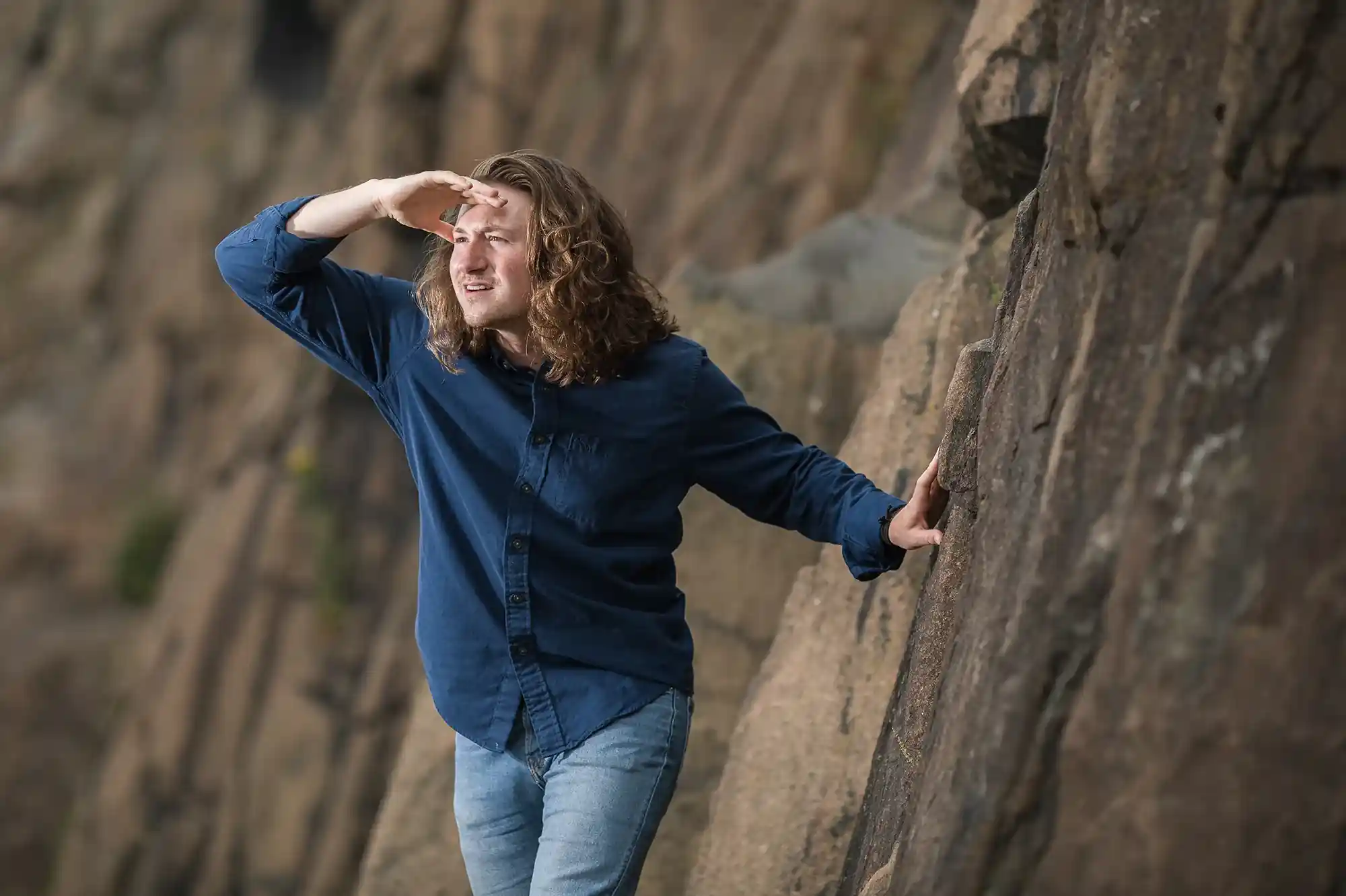 A person with long hair and wearing a blue shirt and jeans uses one hand to shield their eyes while holding onto a rock face with the other hand.